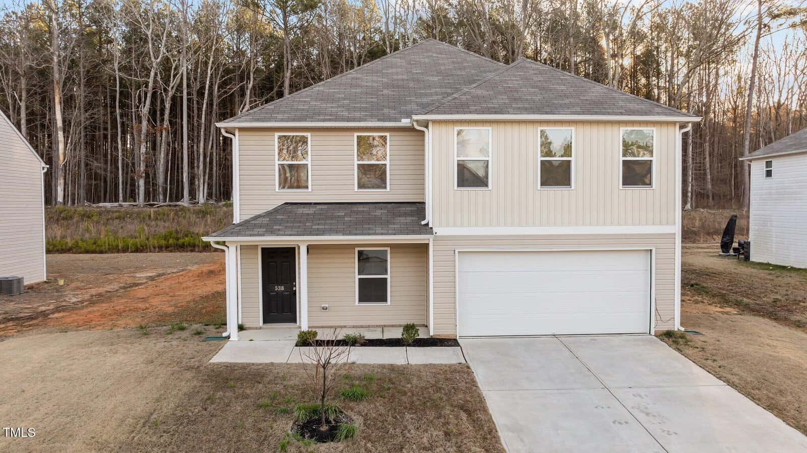 A two-story beige house with dark gray roof shingles and a white two-car garage. The house has a gray front door and small front yard with minimal landscaping. Surrounding the house are areas of dry grass and forested backdrops.