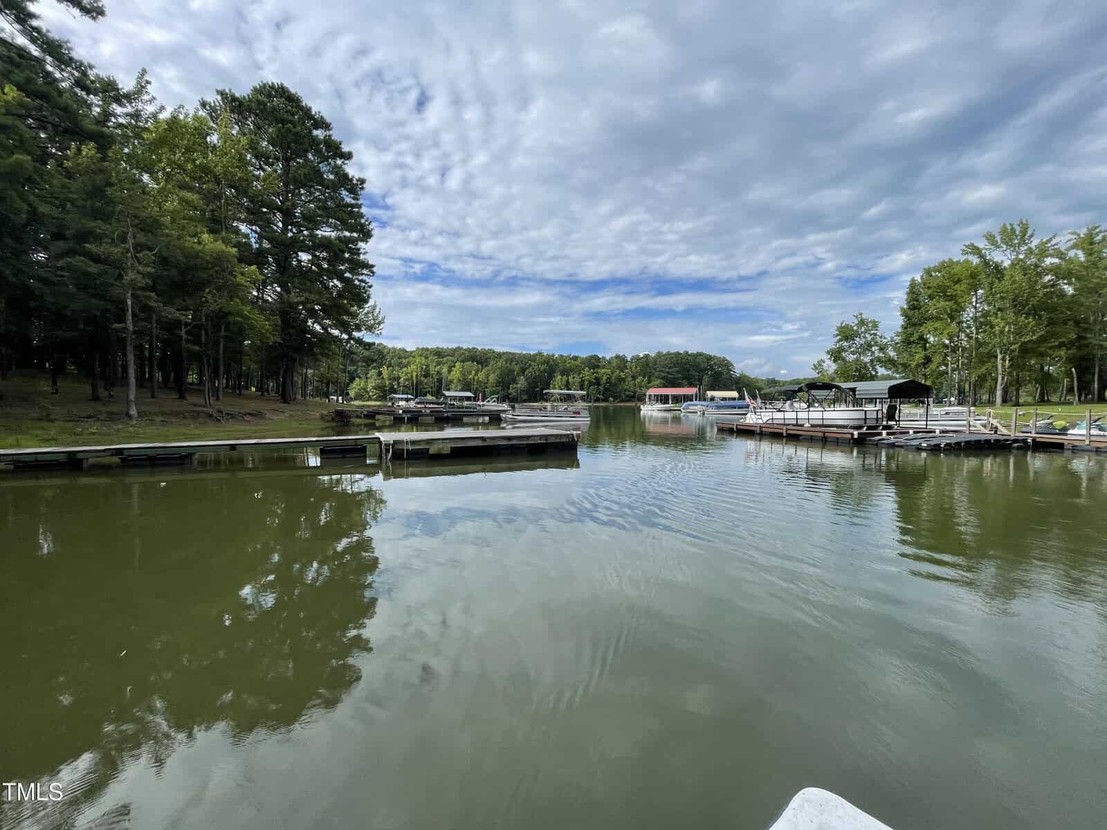 A calm lake scene with several docks and boats moored on either side. The foreground shows still water under a partly cloudy sky. Surrounding the lake are lush green trees and a few structures that seem like boathouses or sheds.