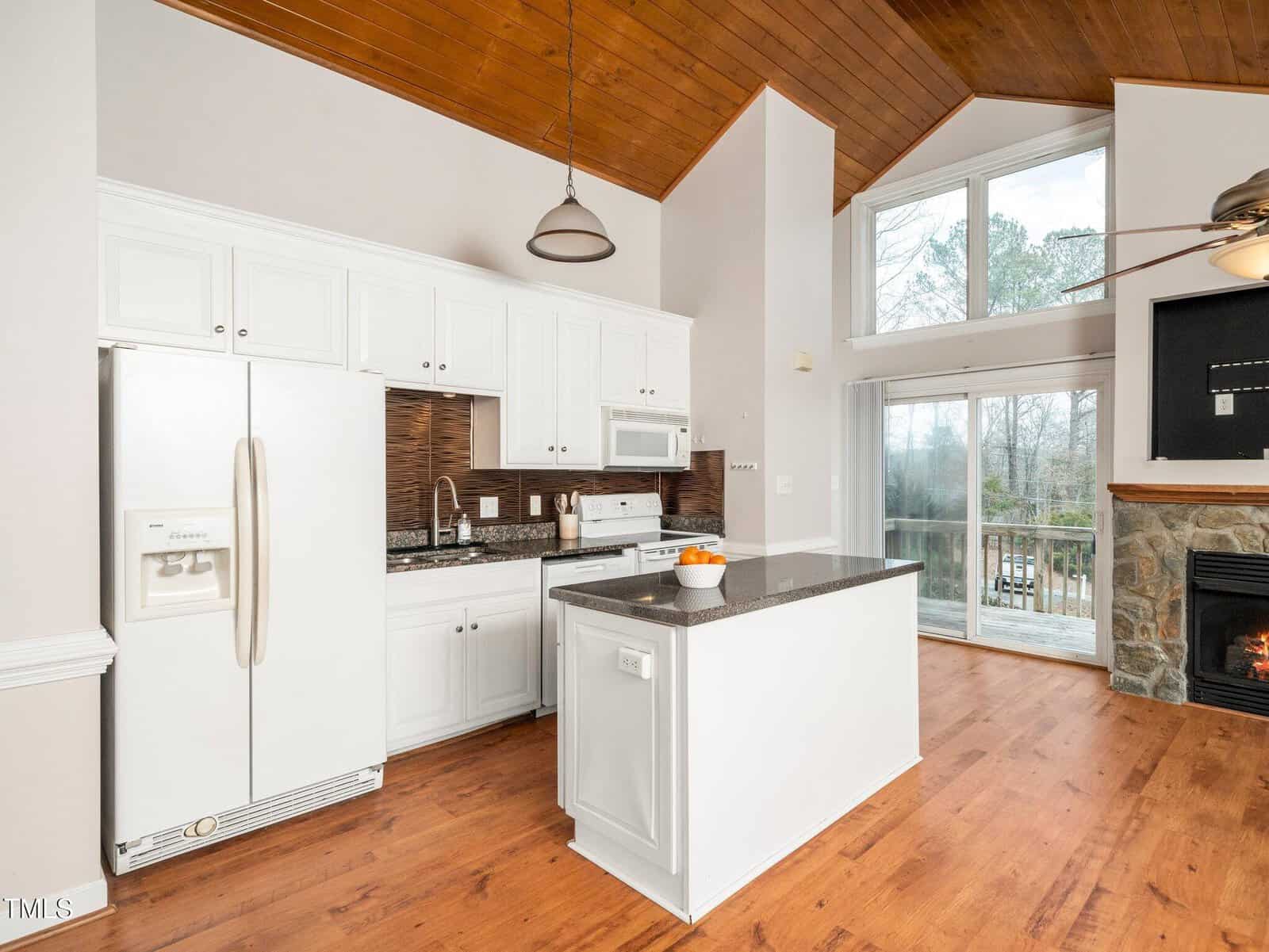 Modern kitchen with white cabinets, granite island, and wood floors. Large windows let in natural light. There is a white refrigerator, oven, microwave, sink, and a stone fireplace in the open living space.