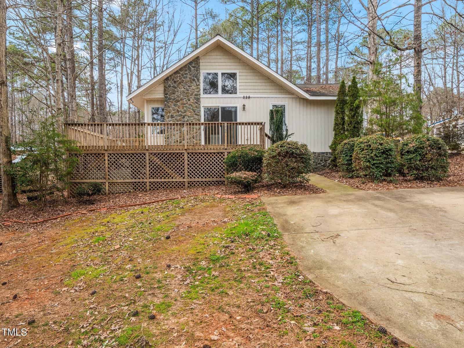 A single-story house with stone and wood siding, large windows, a wooden front deck with a railing, surrounding bushes, and a concrete driveway, set among tall trees with scattered leaves on the ground.