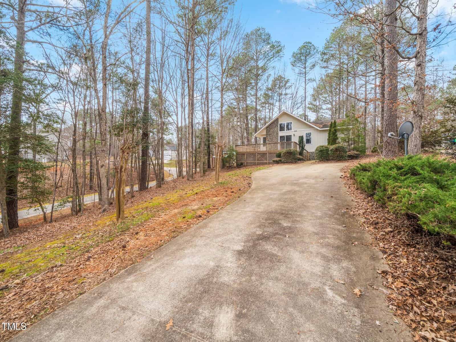 A concrete driveway curves uphill through a wooded area toward a white house with a deck and a satellite dish, surrounded by tall trees and dry leaves on the ground.