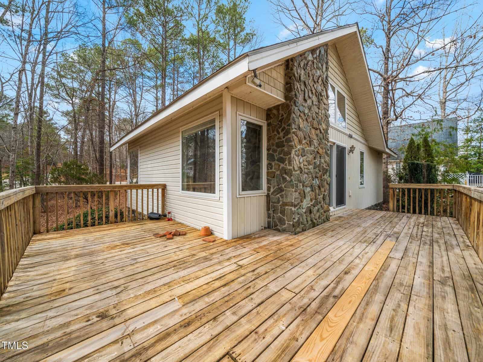 A wooden deck surrounds a modern house with large windows and a stone accent wall. The area is bordered by trees, and the sky is clear with a few clouds. Several empty planters are on the deck.