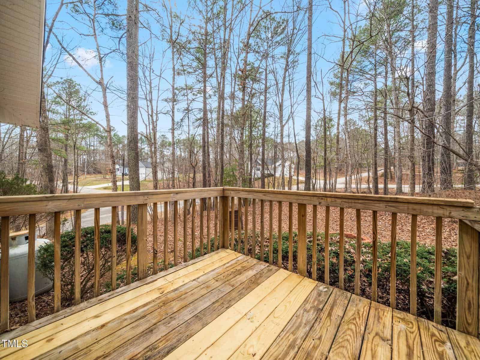 A wooden deck with a simple railing overlooks a yard with leaf-covered ground and tall trees. A road and more trees are visible in the background under a blue sky with some clouds.
