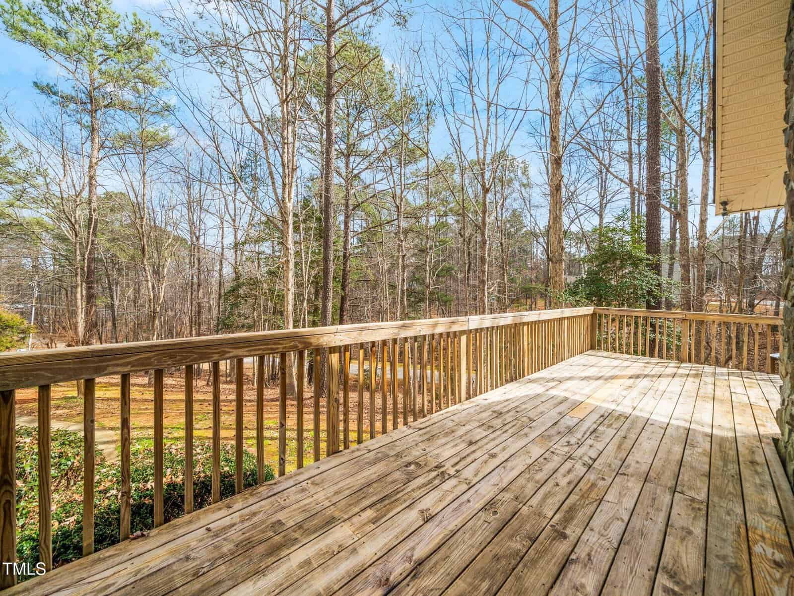 A wooden deck with railings overlooks a wooded backyard with trees and sparse greenery on a clear day. The deck boards are weathered, and there is a small patch of landscape visible below.