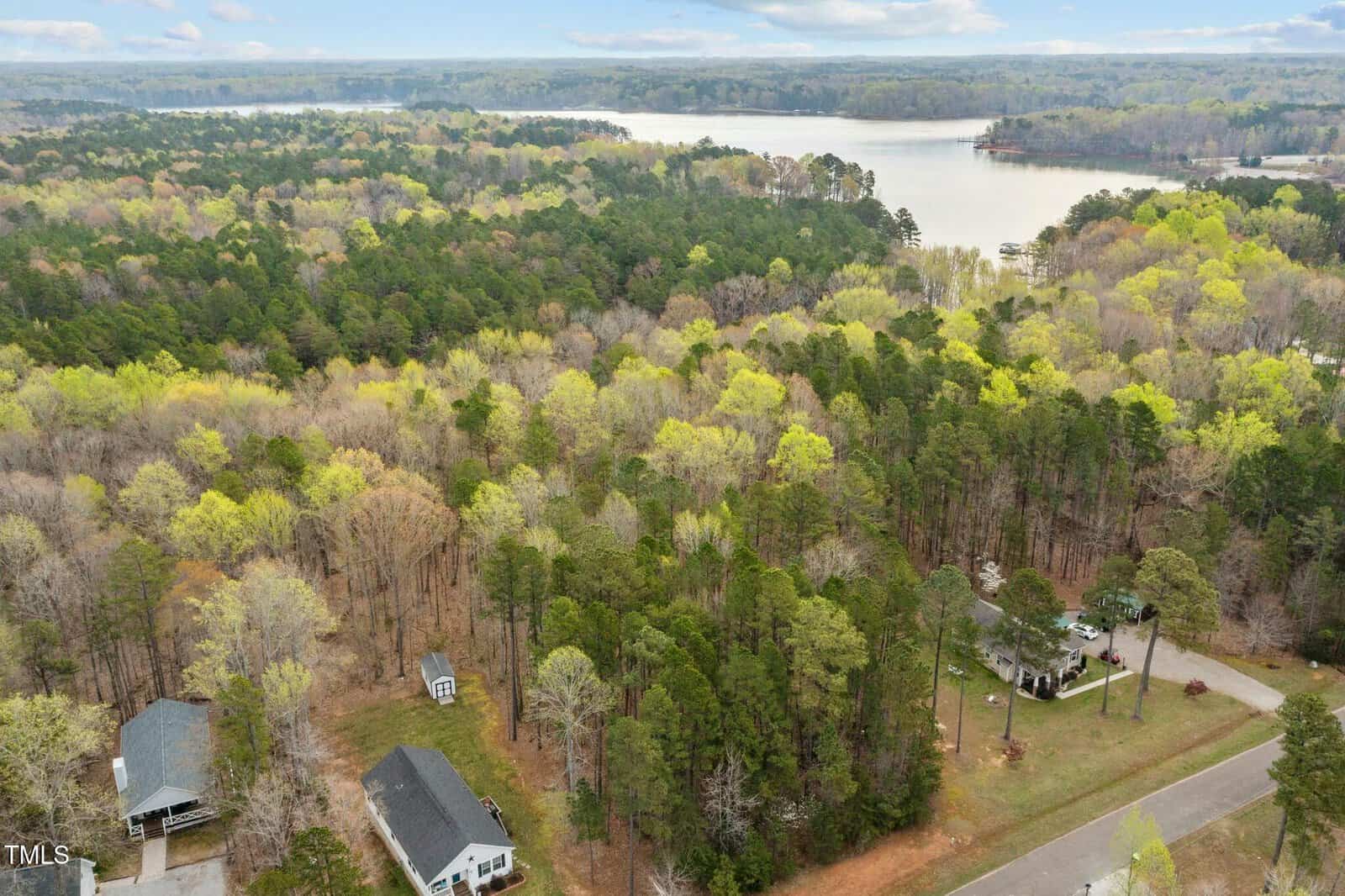 Aerial view of a wooded area with scattered houses and a road. A large body of water is visible in the background. The trees are a mix of evergreen and deciduous, with a few buildings nestled among them. The sky is partly cloudy.