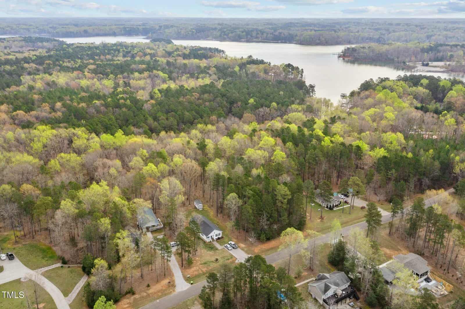 Aerial view of a forested residential area near a lake. Several houses are visible among the trees, with a road running through the neighborhood. The lake is in the background, surrounded by dense woods.