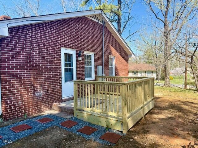 A red brick house with a white door and small wooden deck in the backyard. The ground is bare with scattered leaves and a short path of red bricks. Trees without leaves are visible in the background.