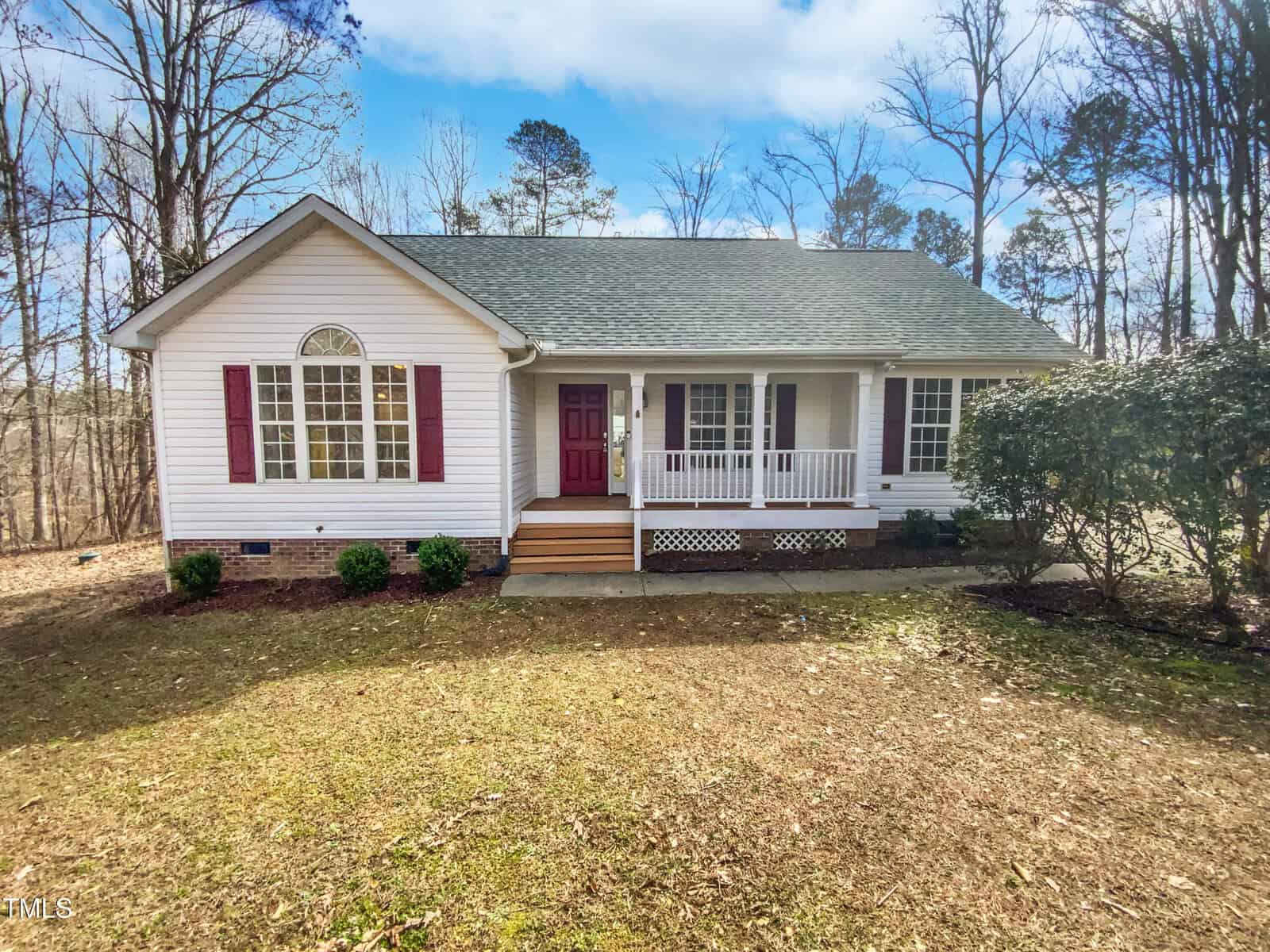 A single-story white house with red shutters, a covered front porch, a red door, and a gable roof, surrounded by trees and a lawn with sparse grass.