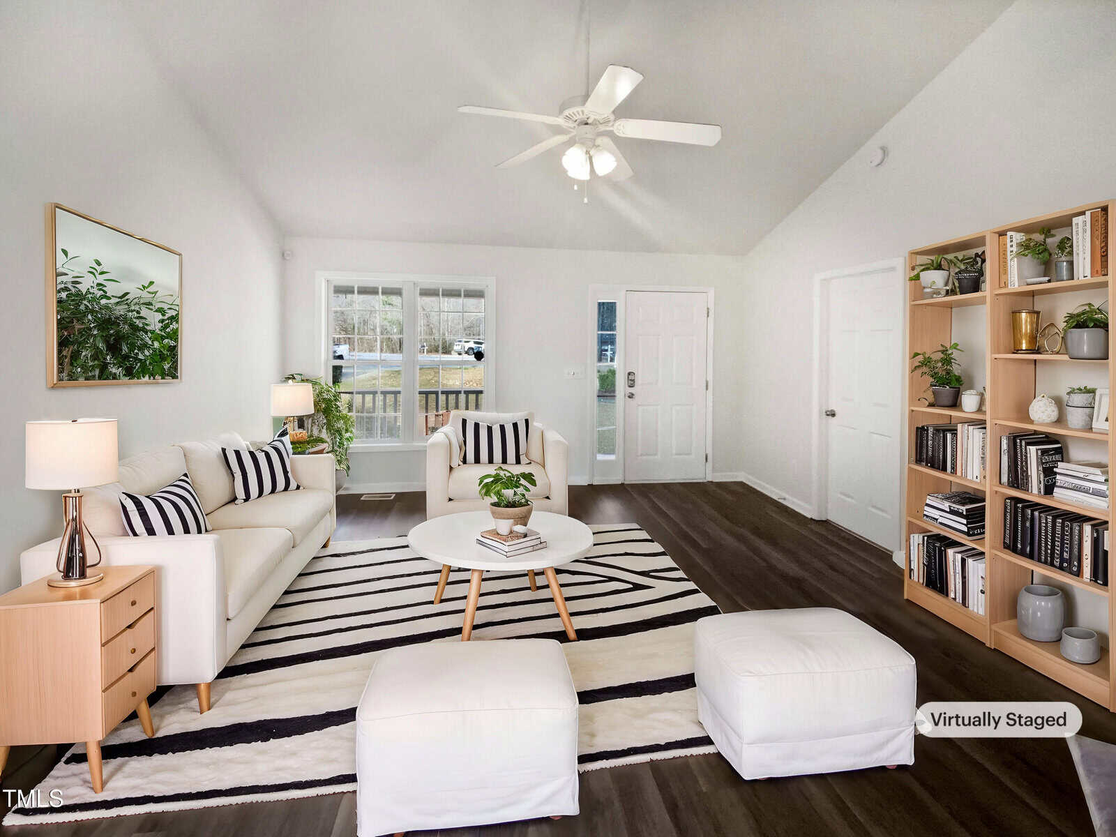 A bright living room with white walls, dark wood flooring, and a ceiling fan. The room has a striped rug, two white ottomans, a coffee table, an armchair, a sofa, a bookshelf, and a side table with a lamp.