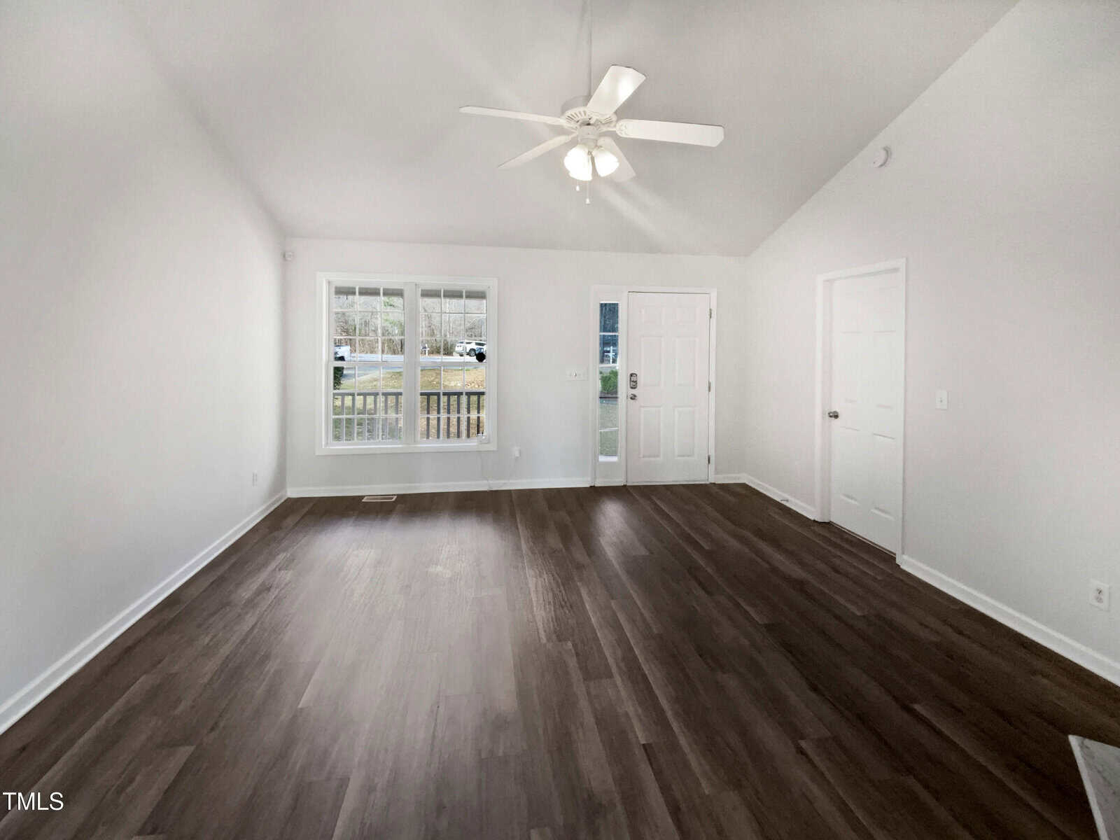 Empty living room with white walls, dark wood flooring, a ceiling fan with lights, large front window, and a white entry door. Natural light enters from the window. The room has a high, sloped ceiling.