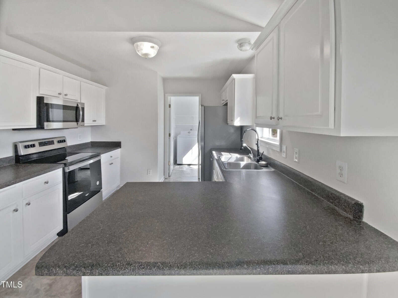 A kitchen with white cabinets, black countertops, stainless steel appliances, a double sink, and an overhead light fixture. A doorway at the back leads to another room with a window.