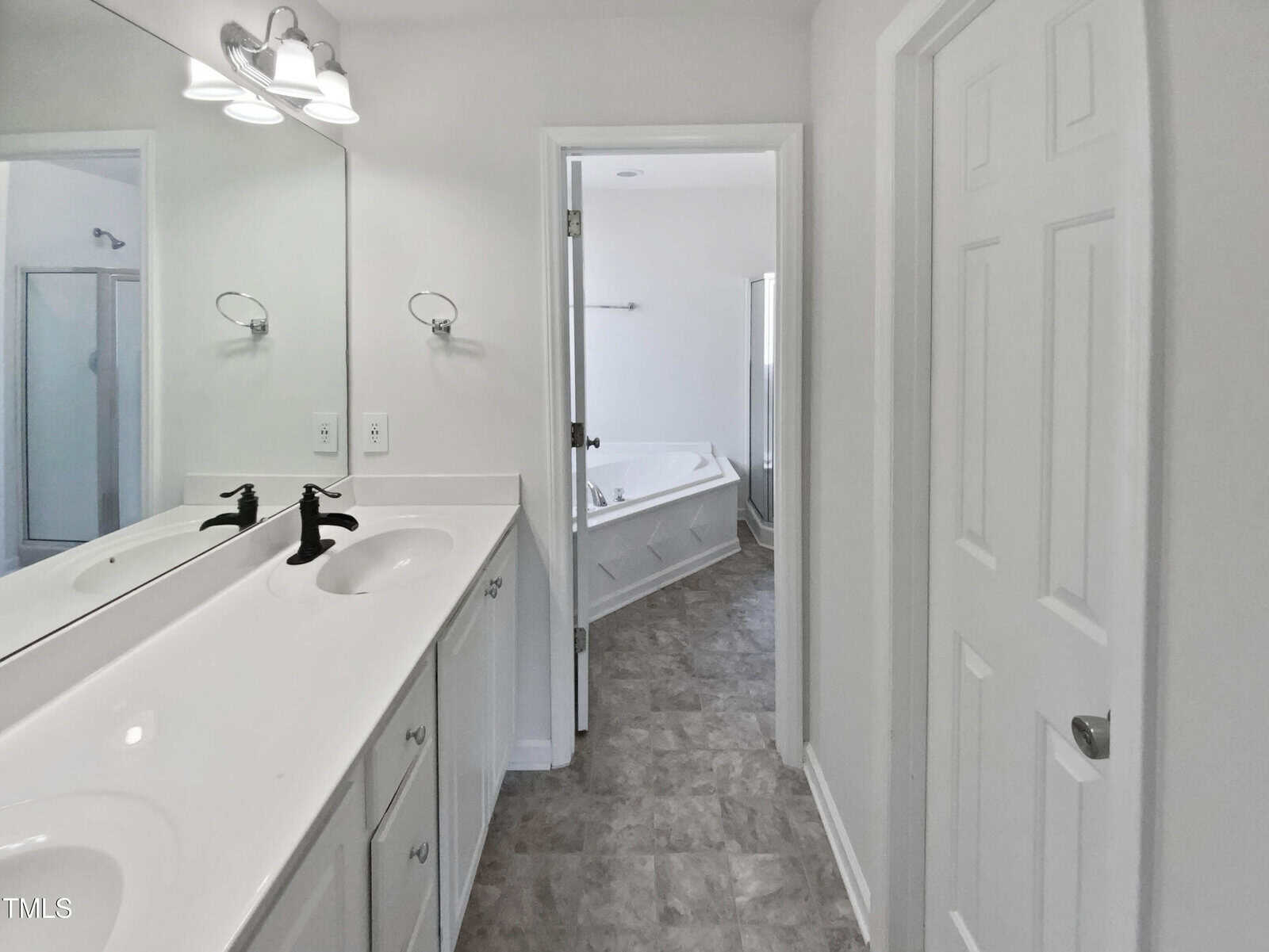 White bathroom with double sinks, large mirror, black faucets, gray floor tiles, towel ring, and a doorway leading to a separate bathtub and shower area with a white door partially open.