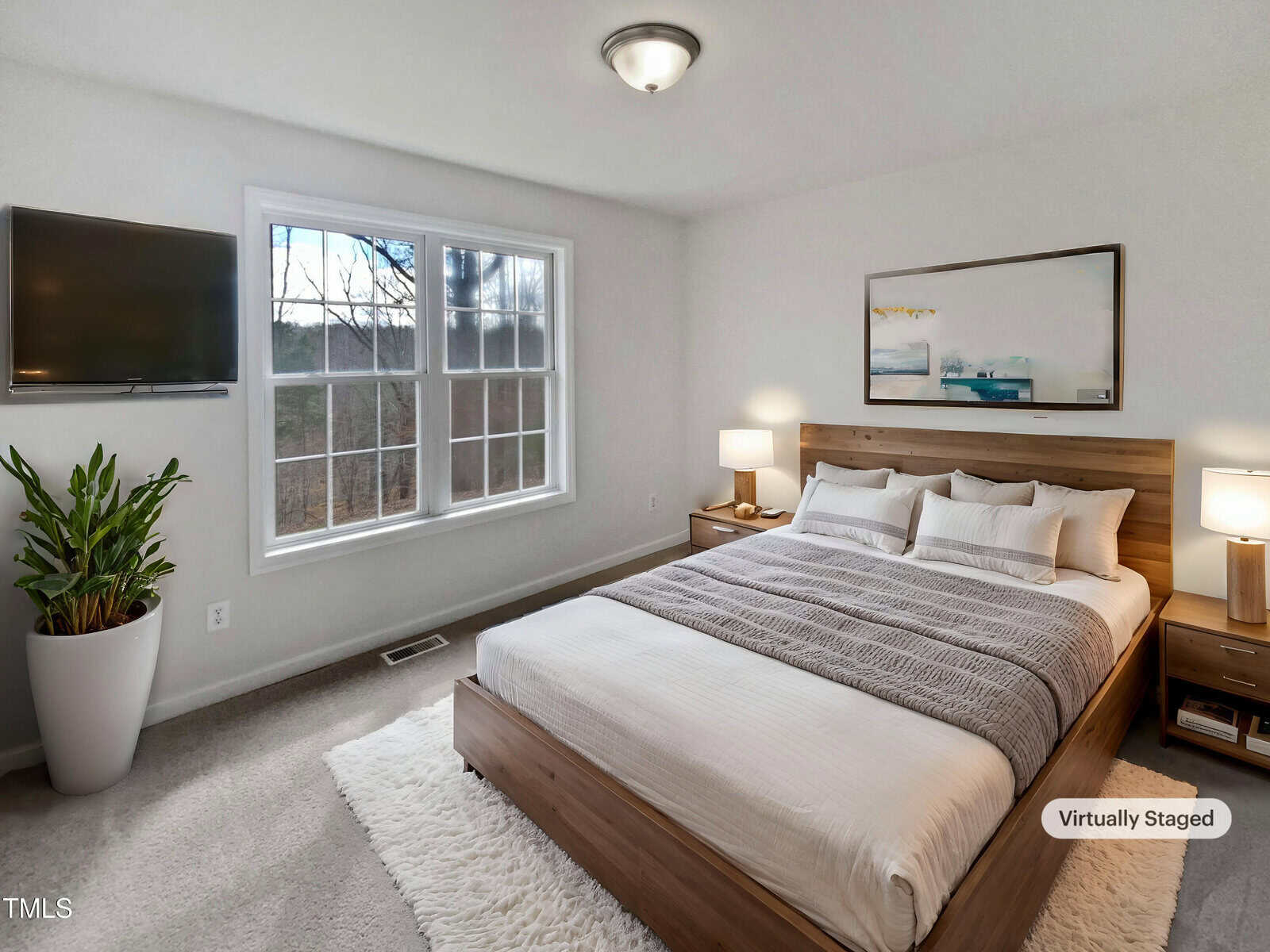A bedroom with a large bed featuring white and gray bedding, two matching nightstands with lamps, a wall-mounted TV, a potted plant, a rug, and a large window letting in natural light. A "Virtually Staged" label is visible.