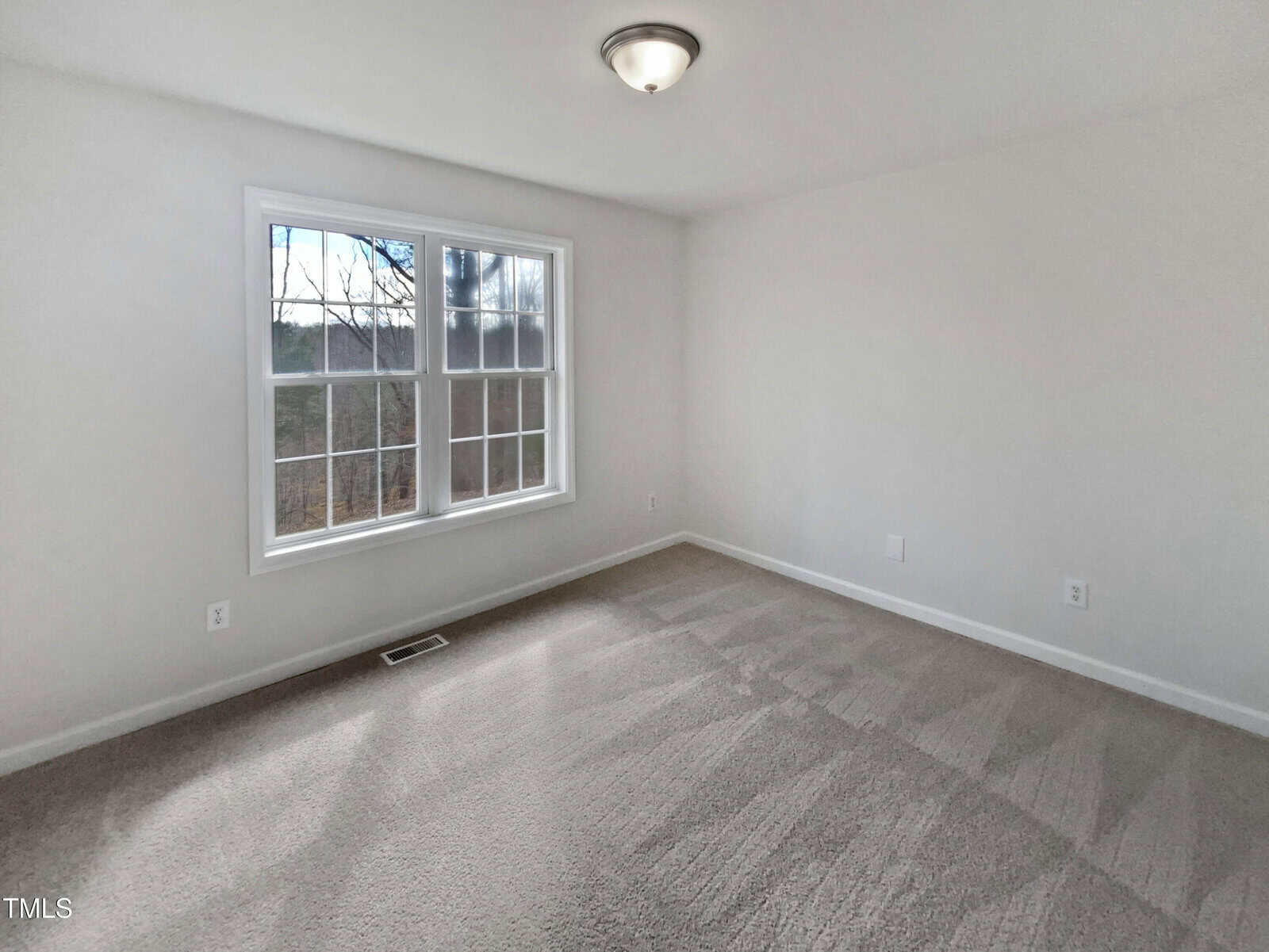 Empty room with beige carpet, white walls, a large double window letting in natural light, and a ceiling light fixture. Electrical outlets are visible along the base of the walls.
