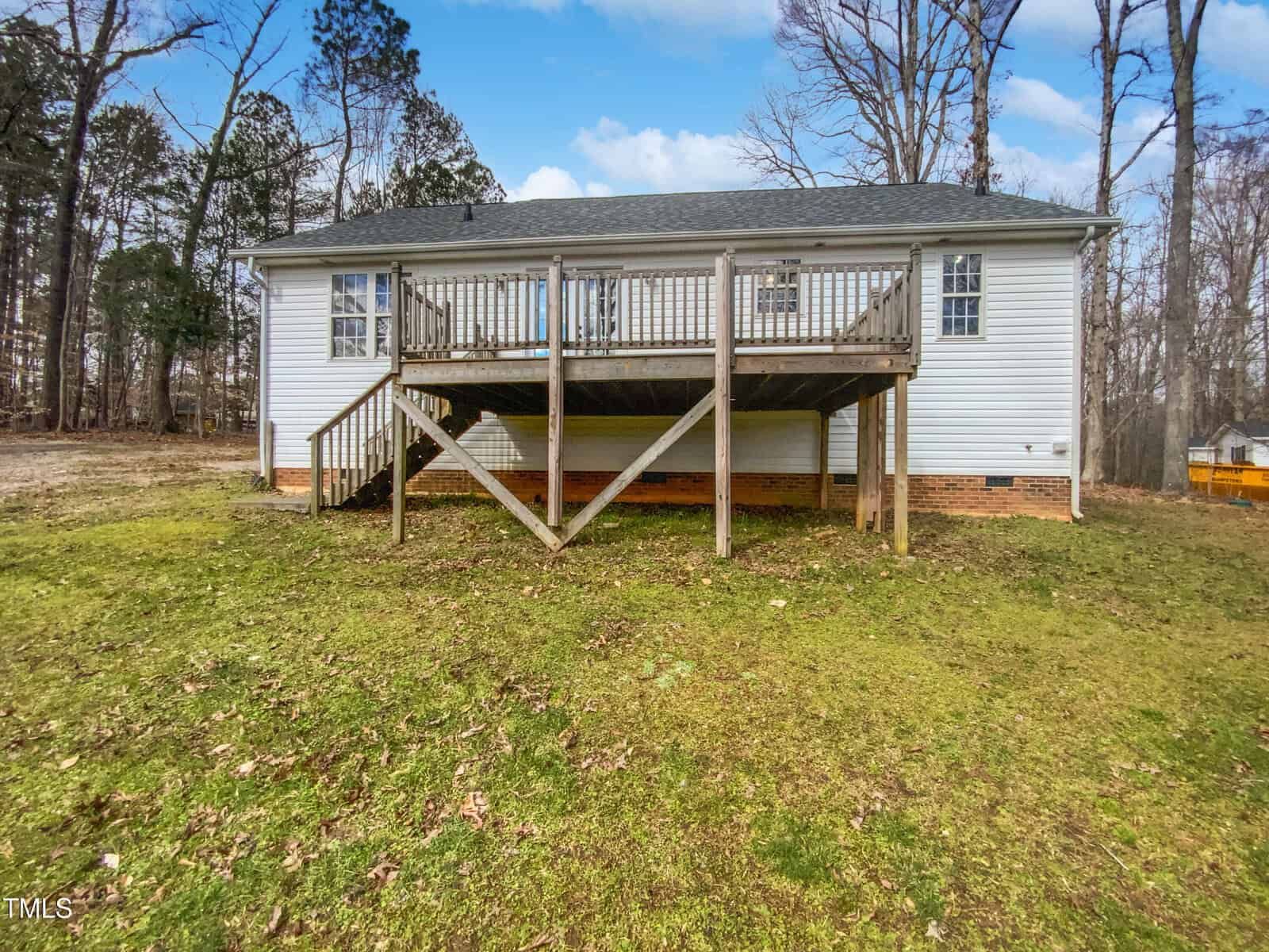 A white single-story house with a large wooden deck and stairs in the backyard, surrounded by leafless trees and patchy grass on the ground under a partly cloudy sky.