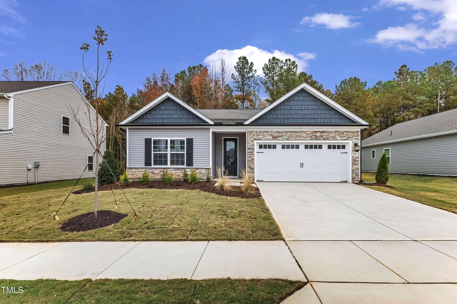Single-story suburban house with gray siding, stone accents, and a double garage. A concrete driveway and walkway lead to the front door. The lawn is newly planted, and neighboring homes are visible on each side.