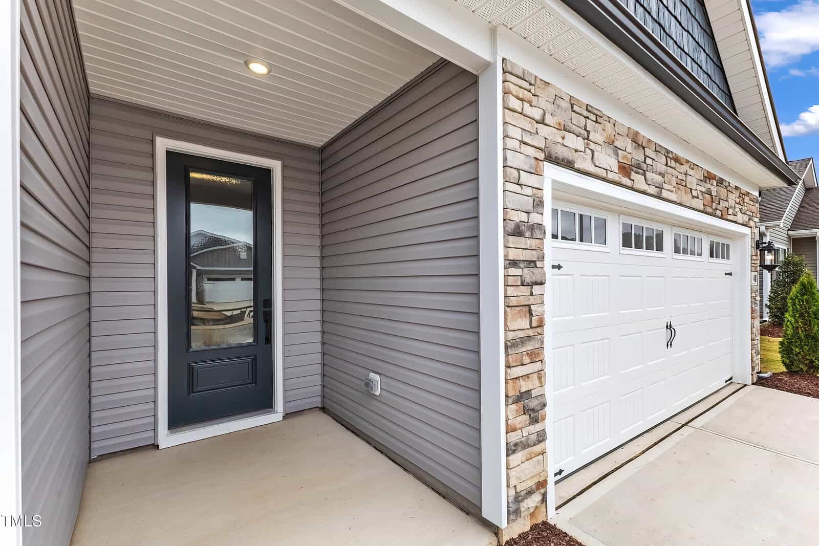 A house exterior showing a covered entryway with a dark framed glass front door, adjacent to a white double garage door with windows, stone and gray siding, and a concrete walkway.