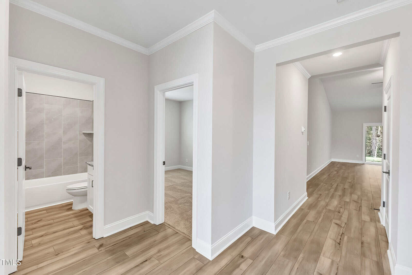 A hallway with light wood flooring leads to a carpeted room, a bathroom with a bathtub and toilet, and another area at the end. The walls are light gray with white trim and crown molding.
