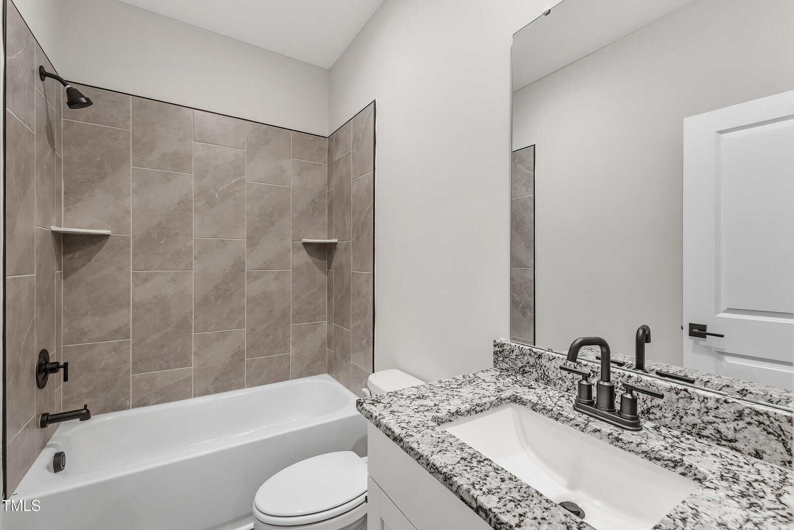A modern bathroom with a bathtub-shower combo featuring beige tiles, a white toilet, and a granite countertop with a rectangular sink. The fixtures are matte black, and the walls are painted white. A mirror hangs above the sink.
