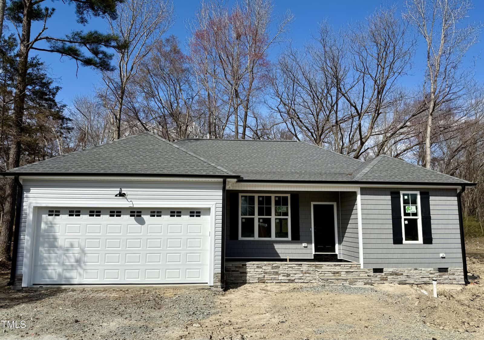 Located at 600 Hicksmill Road in Oxford, a single-story gray house with a gabled roof is under construction. It boasts a large garage door on the left and a quaint porch on the right. The exterior has stone accents, and it's surrounded by leafless trees under a clear blue sky.
