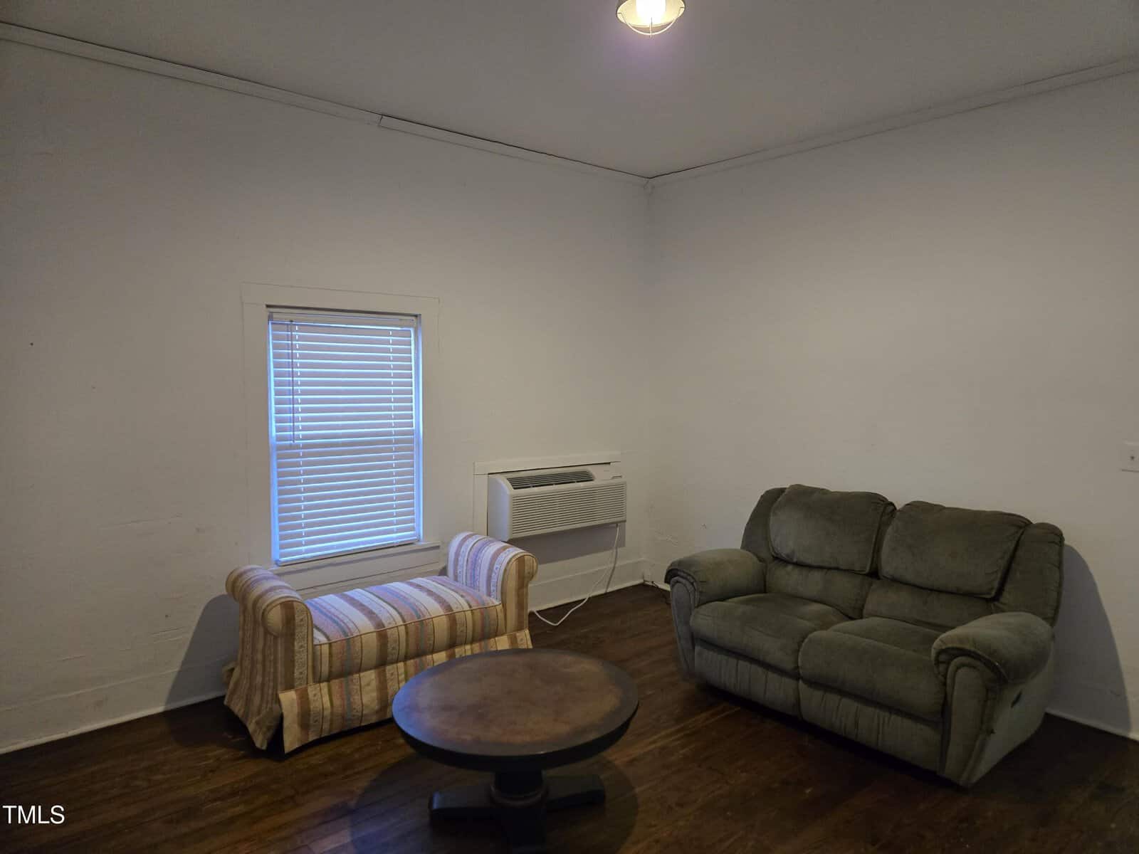 A small room with a striped chaise lounge, a green loveseat, a round wooden coffee table, an air conditioning unit below a window with blinds, white walls, and dark wood flooring.