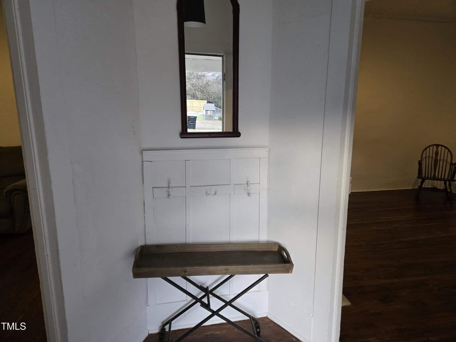 A small wooden bench with a metal base sits in a white alcove beneath a wall mirror. Coat hooks are mounted on the wall. Part of a separate room with hardwood floors and a wooden chair is visible.