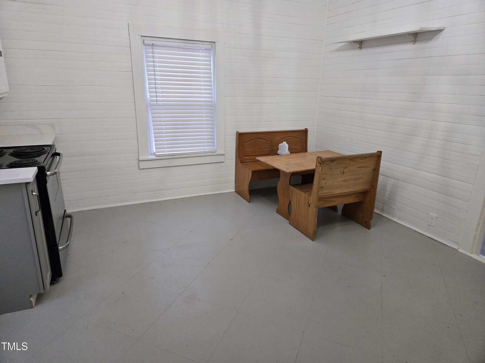 A small kitchen with a window with blinds, a stove and oven on the left, and a wooden dining nook with bench seating and a rectangular table in the corner. A paper towel roll sits on the table.