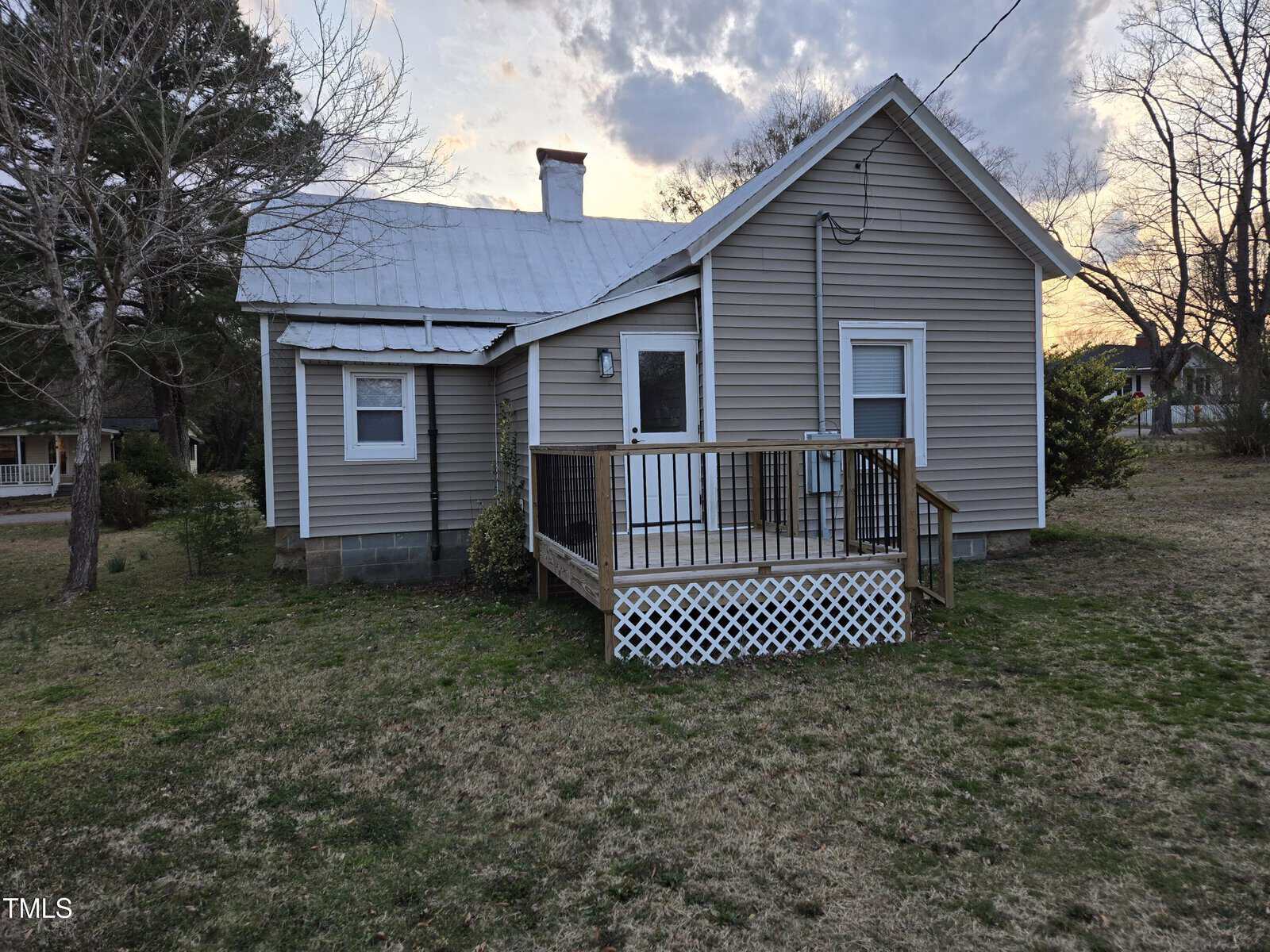 A small beige house with white trim and a metal roof stands on a grassy yard. A wooden porch with a white lattice panel and simple railing is attached to the side, and leafless trees are in the background under a cloudy sky.