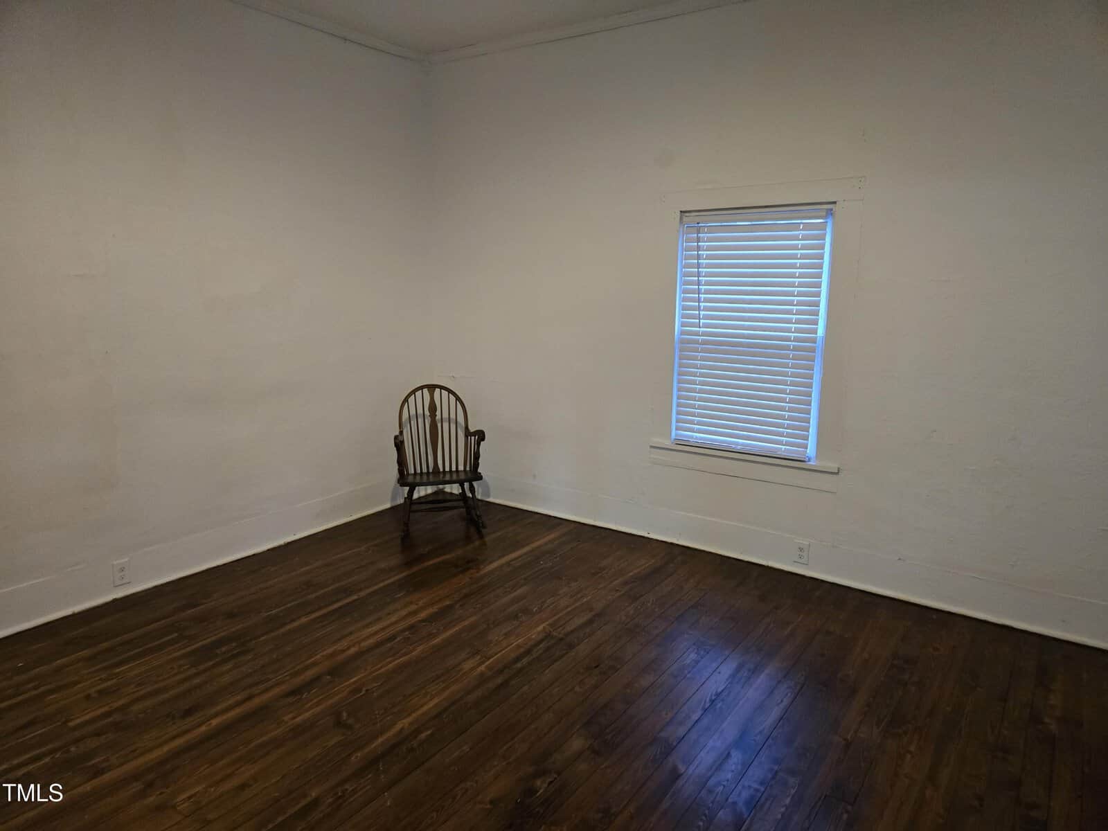 A mostly empty room with white walls, dark wood floors, a single wooden chair in the corner, and one window with closed blinds.