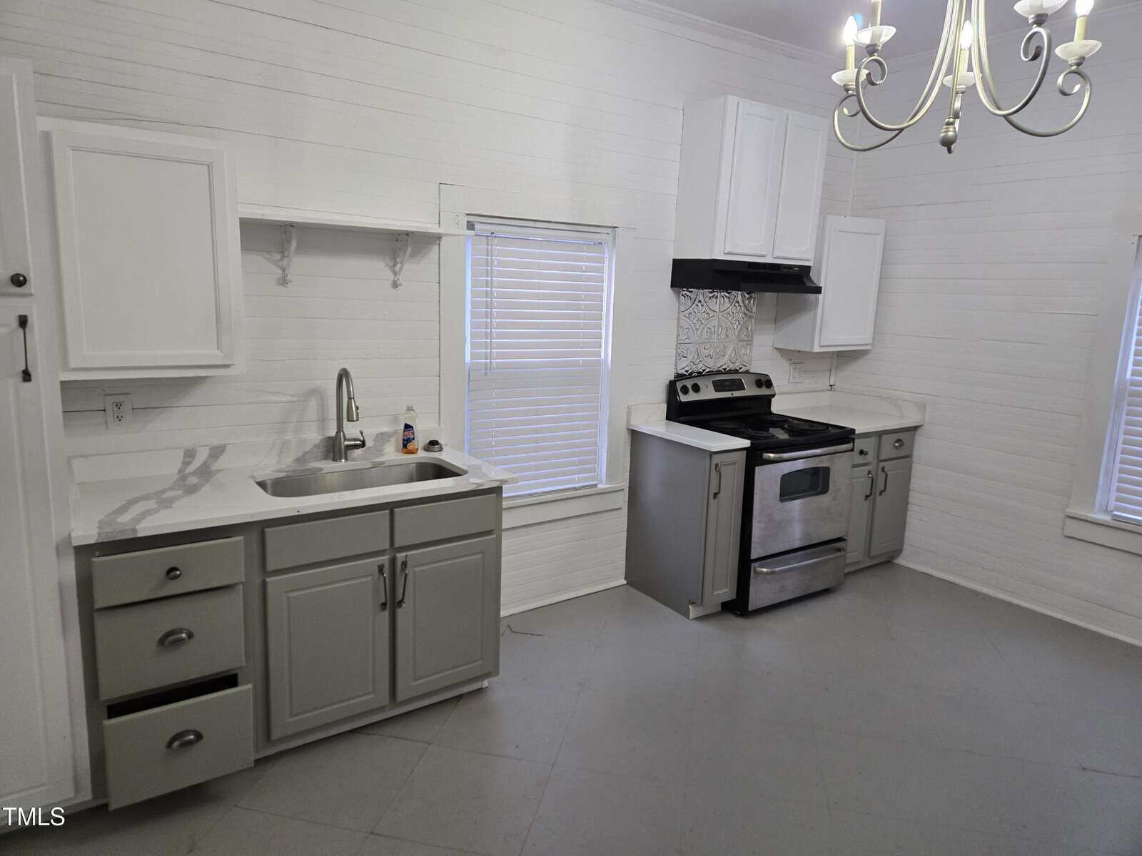 A kitchen with light gray lower cabinets, white upper cabinets, a sink, an electric stove with an oven, a vent hood, white blinds on two windows, and a white chandelier hanging from the ceiling.