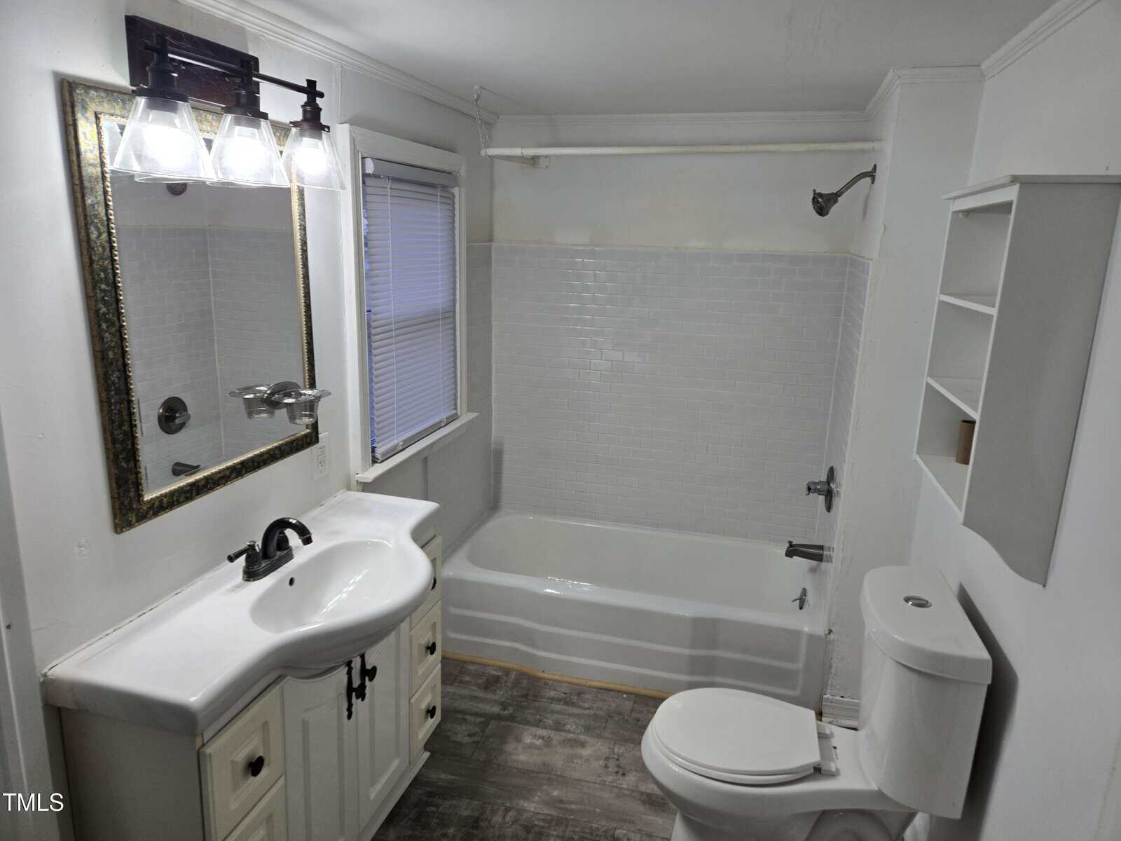 A bathroom with a white sink and vanity, a wall mirror with lights above, a toilet, a bathtub with a shower, tiled walls around the tub, open shelves, and a window with closed blinds; gray wood-look flooring.