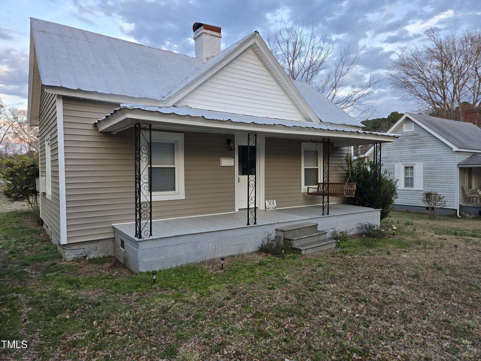 A single-story house with light gray siding, white trim, a metal roof, and a covered front porch featuring a wooden swing. Concrete steps lead up to the porch, and the yard has patchy grass.