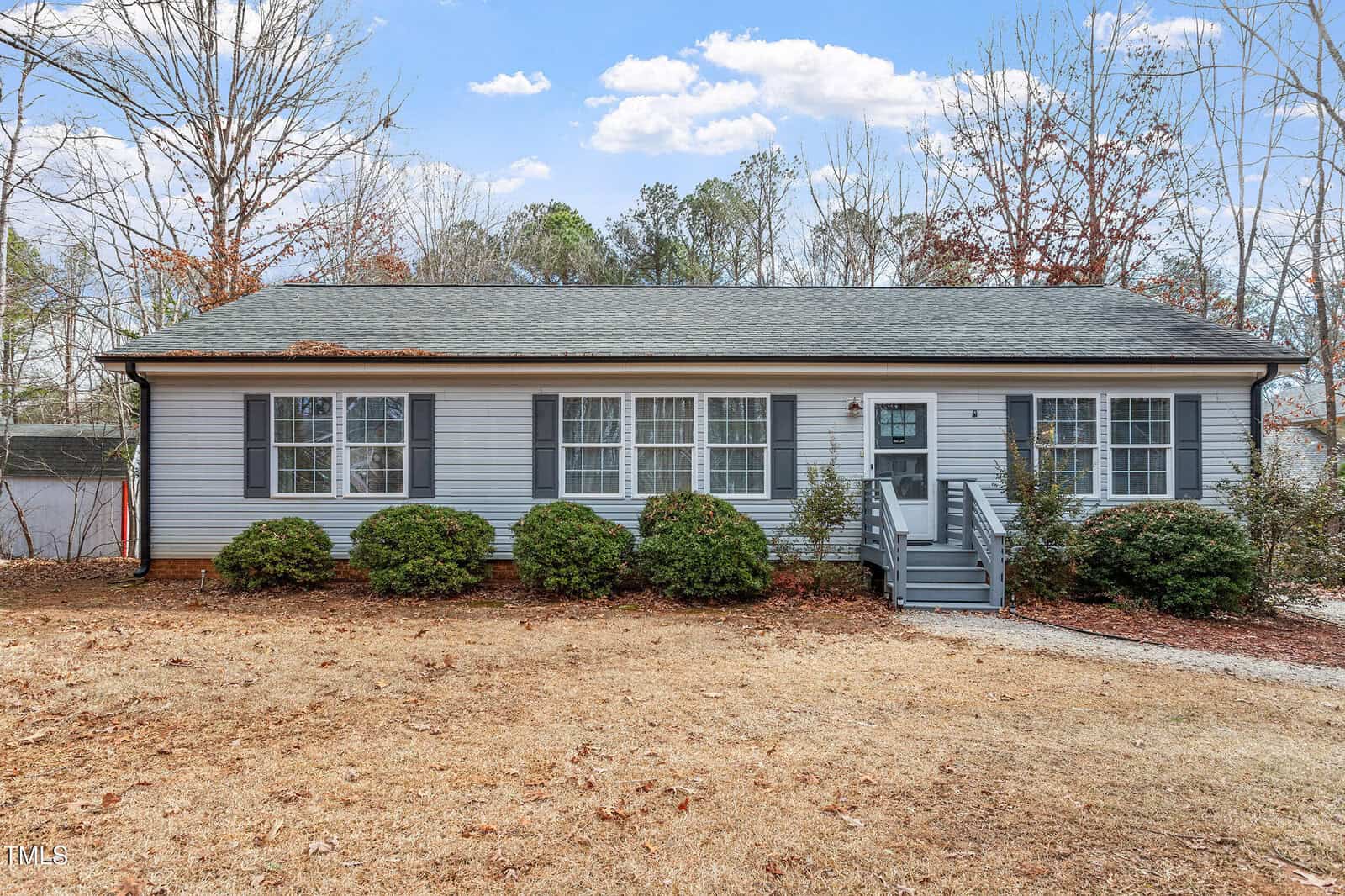 Single-story gray house with white trim, a dark gray roof, and multiple large windows. A small front porch with stairs leads to the entrance. Sparse shrubs and dry grass cover the front yard; trees and a blue sky are in the background.