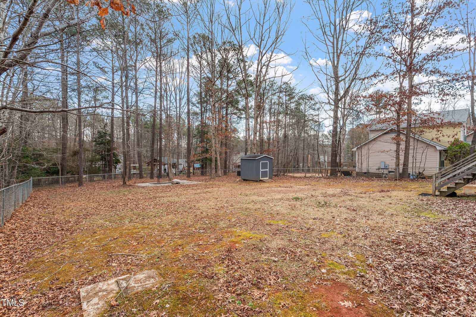A fenced backyard with sparse grass and scattered leaves, featuring a small blue shed, several trees with bare branches, a house on the right, and a few clouds in the blue sky.
