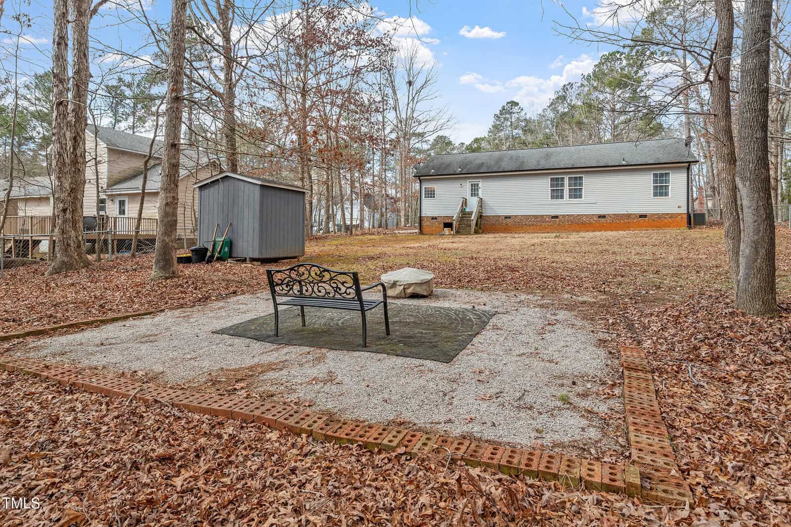 A metal bench sits on a paved area surrounded by gravel and bricks in a backyard. Nearby are a small shed, fallen leaves, leafless trees, and a single-story house with stairs leading to the yard.