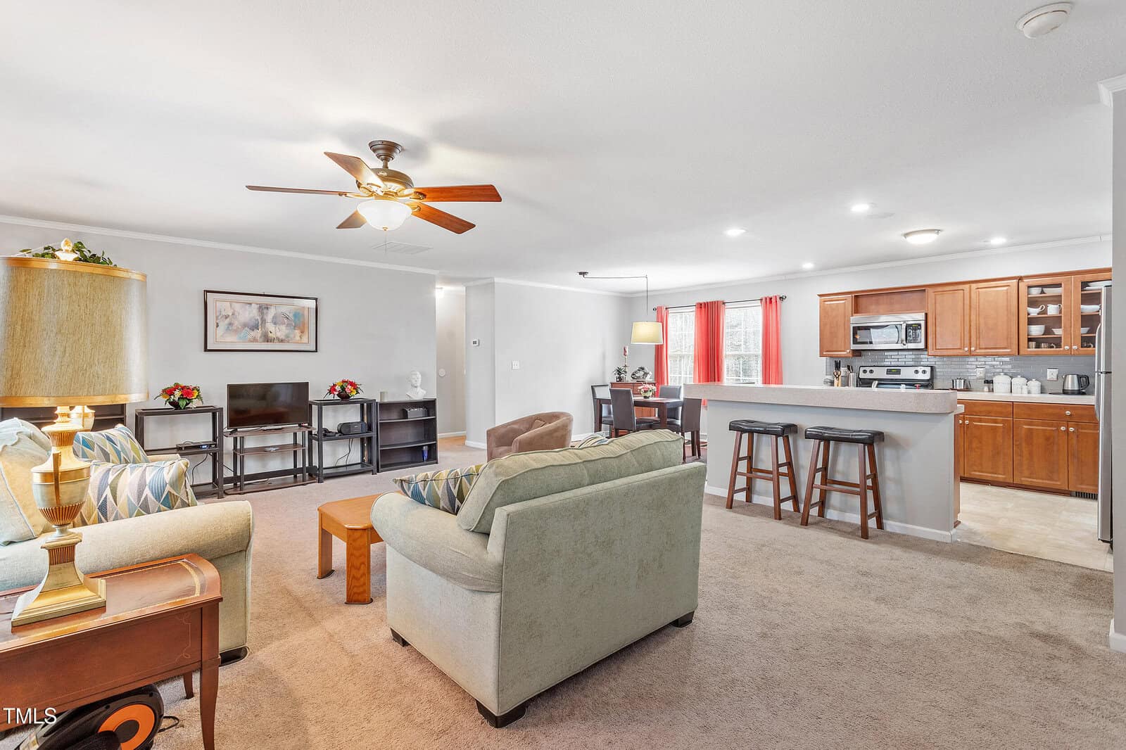 Bright living room with two sofas, an armchair, and a coffee table facing a TV. The open layout includes a kitchen with wooden cabinets, stainless steel appliances, a breakfast bar, and barstools. Red curtains frame a window.