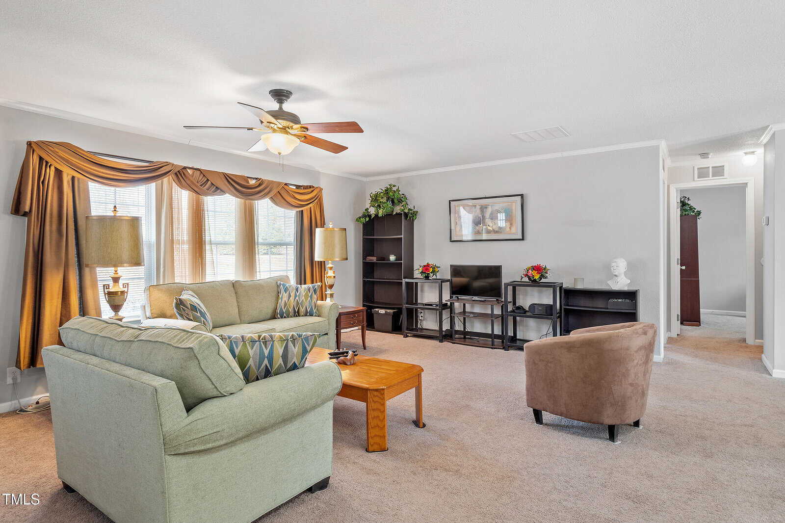 Living room with beige carpet, two sofas, a coffee table, bookshelves, TV, armchair, large windows with drapes, ceiling fan with light, and a hallway leading to another part of the house.