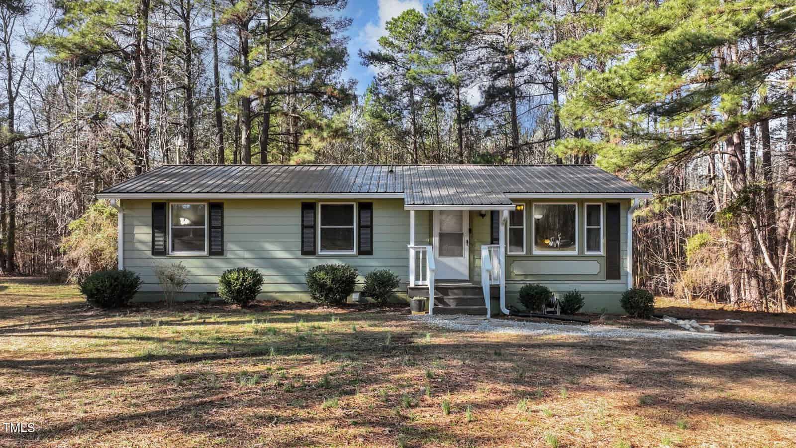 Nestled in a wooded area at 749 Fred Royster Road, Henderson, this small, single-story house features light green siding and a metal roof. A pathway meanders past several bushes to a porch with white railings, all surrounded by tall trees basking in the sun.
