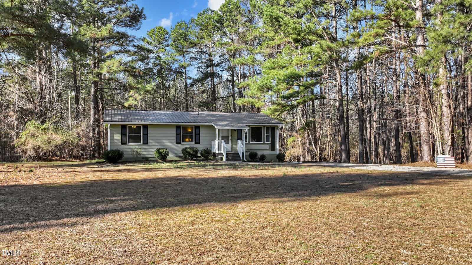 A small single-story house with a gray exterior and white trim sits in a clearing surrounded by tall trees. The lawn is sparse with brown grass, and a gravel driveway leads up to the entrance. The sky is clear with a few clouds.
