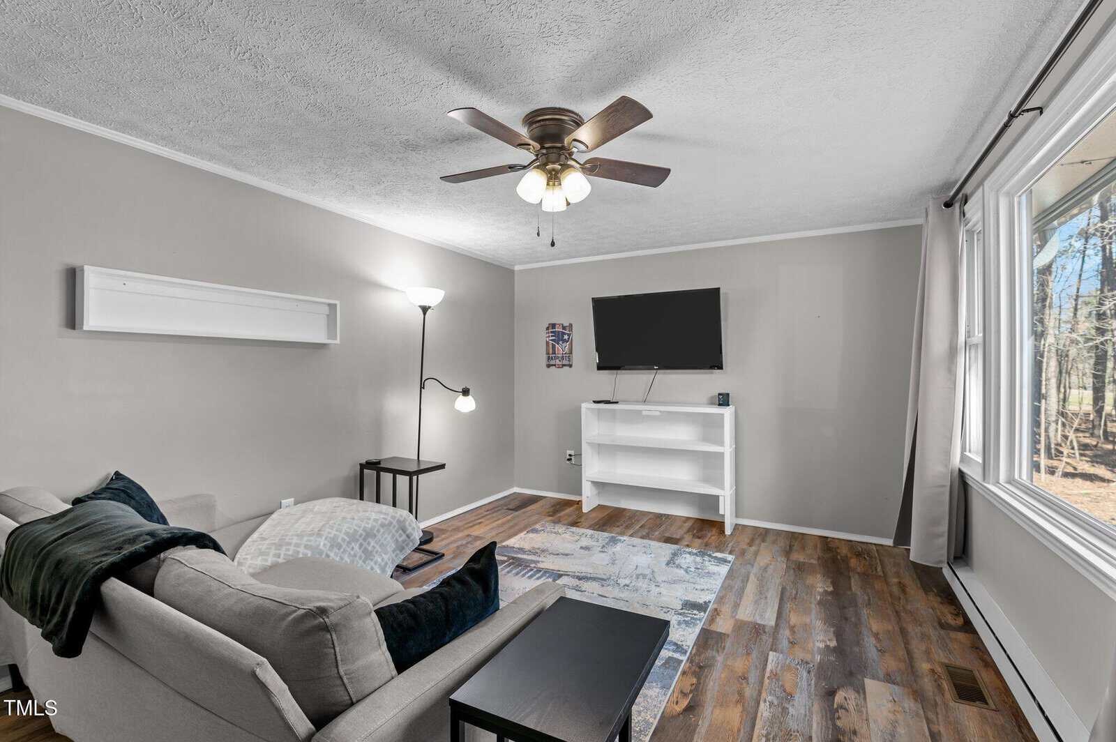 A living room with a gray sofa, black side table, and a freestanding lamp. The walls are light gray, and the floor is wooden. A flat-screen TV sits on a white stand. A ceiling fan with lights is above, and a window lets in natural light.
