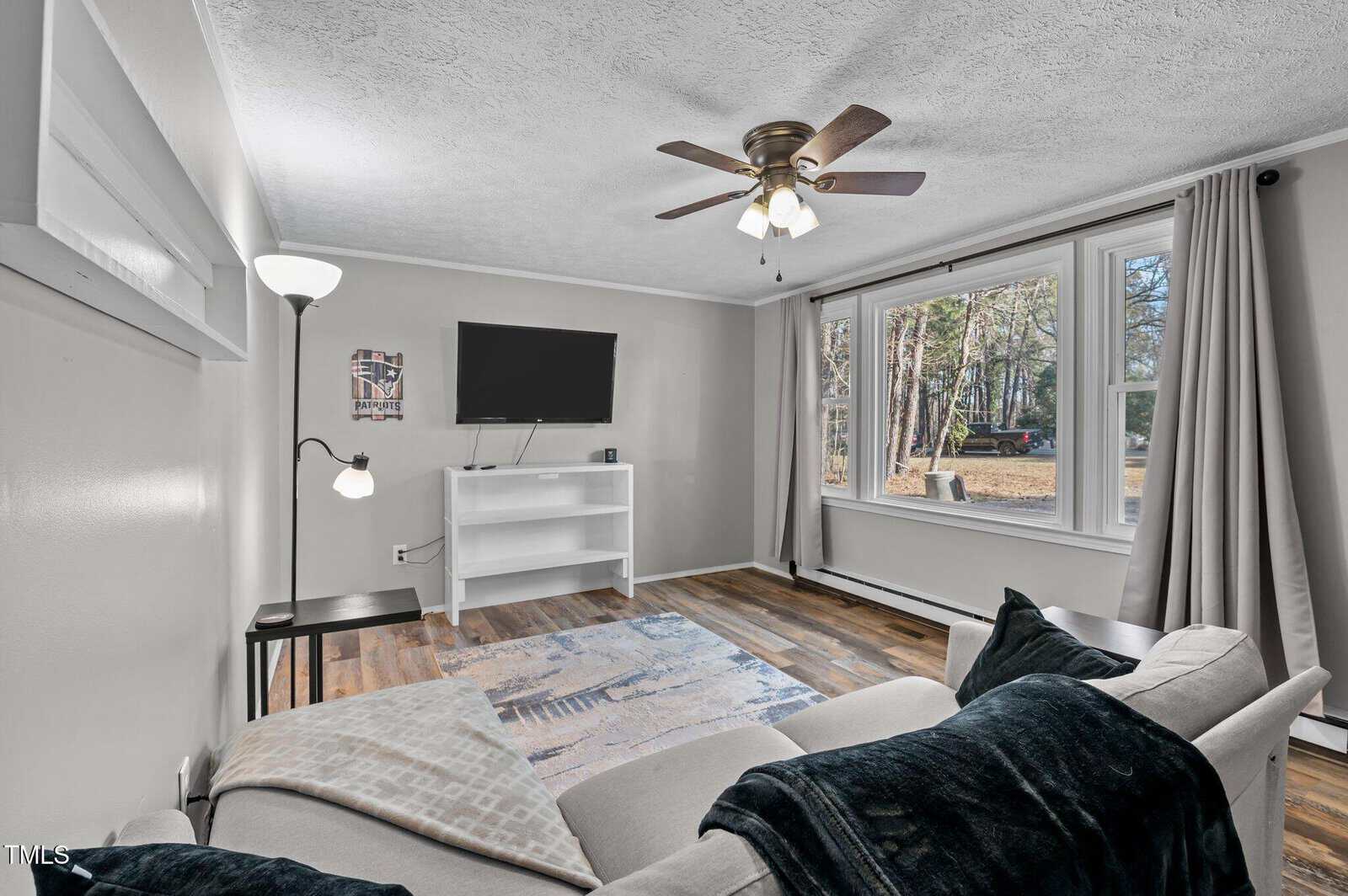 Living room with a ceiling fan, wall-mounted TV, and a white media console. A sofa with cushions faces the TV, and a floor lamp stands nearby. Large windows reveal a view of trees outside. Hardwood floor and soft-colored walls complete the scene.