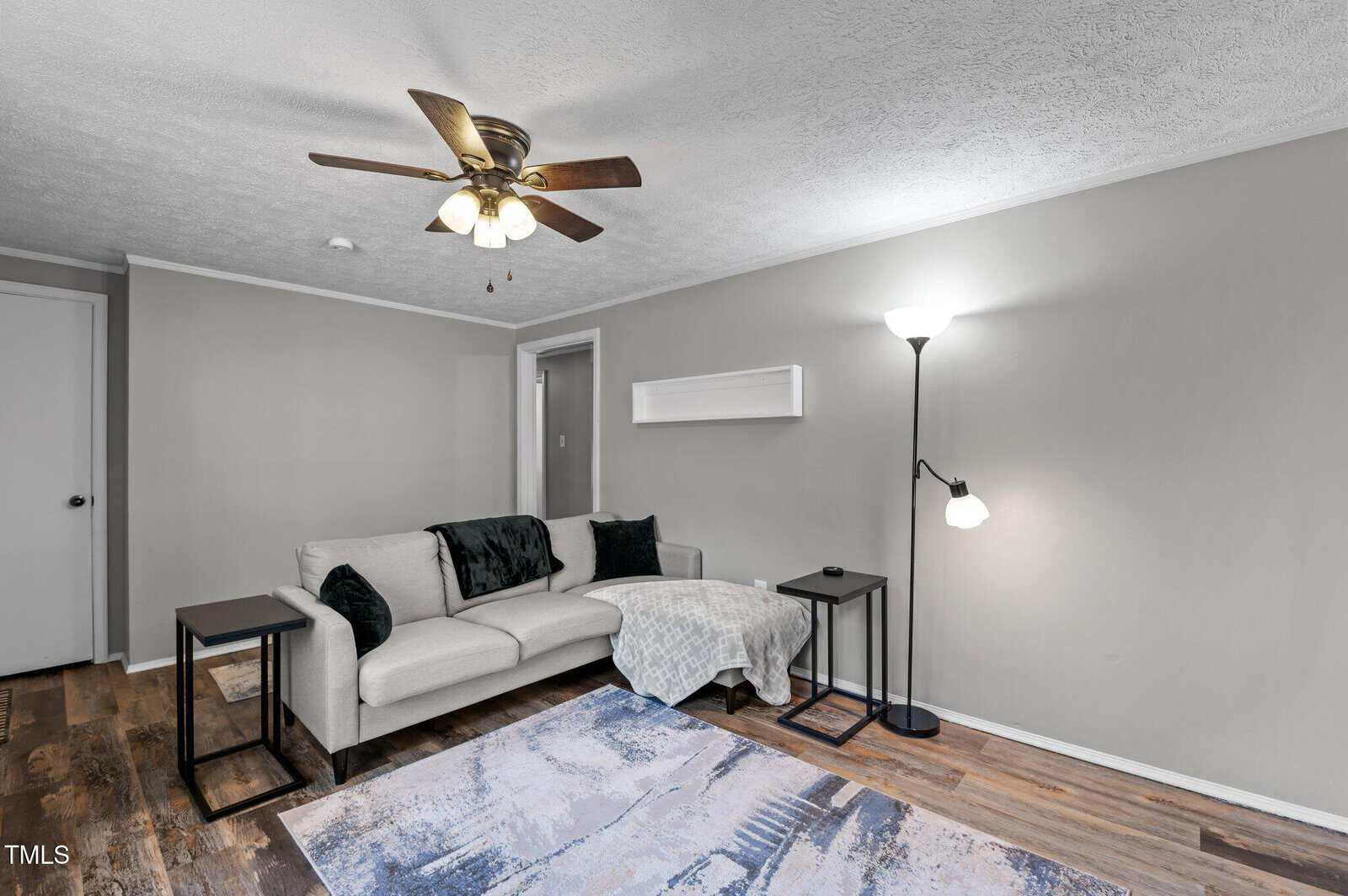 A living room with a beige sectional sofa and dark cushions, a patterned rug on wooden flooring, a floor lamp, and an overhead ceiling fan. Walls are painted light gray with a white door visible in the background.