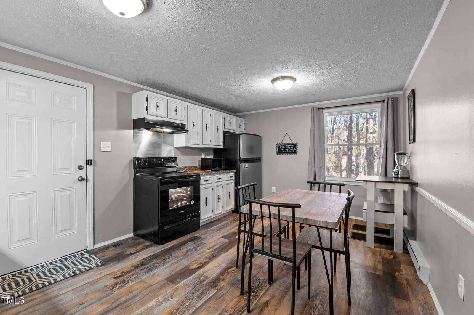 A small kitchen with white cabinets, black appliances, and a wooden dining table with four chairs. A window to the right has a view of trees outside. The floor is wood, and the walls are painted light gray. A light fixture hangs from the ceiling.