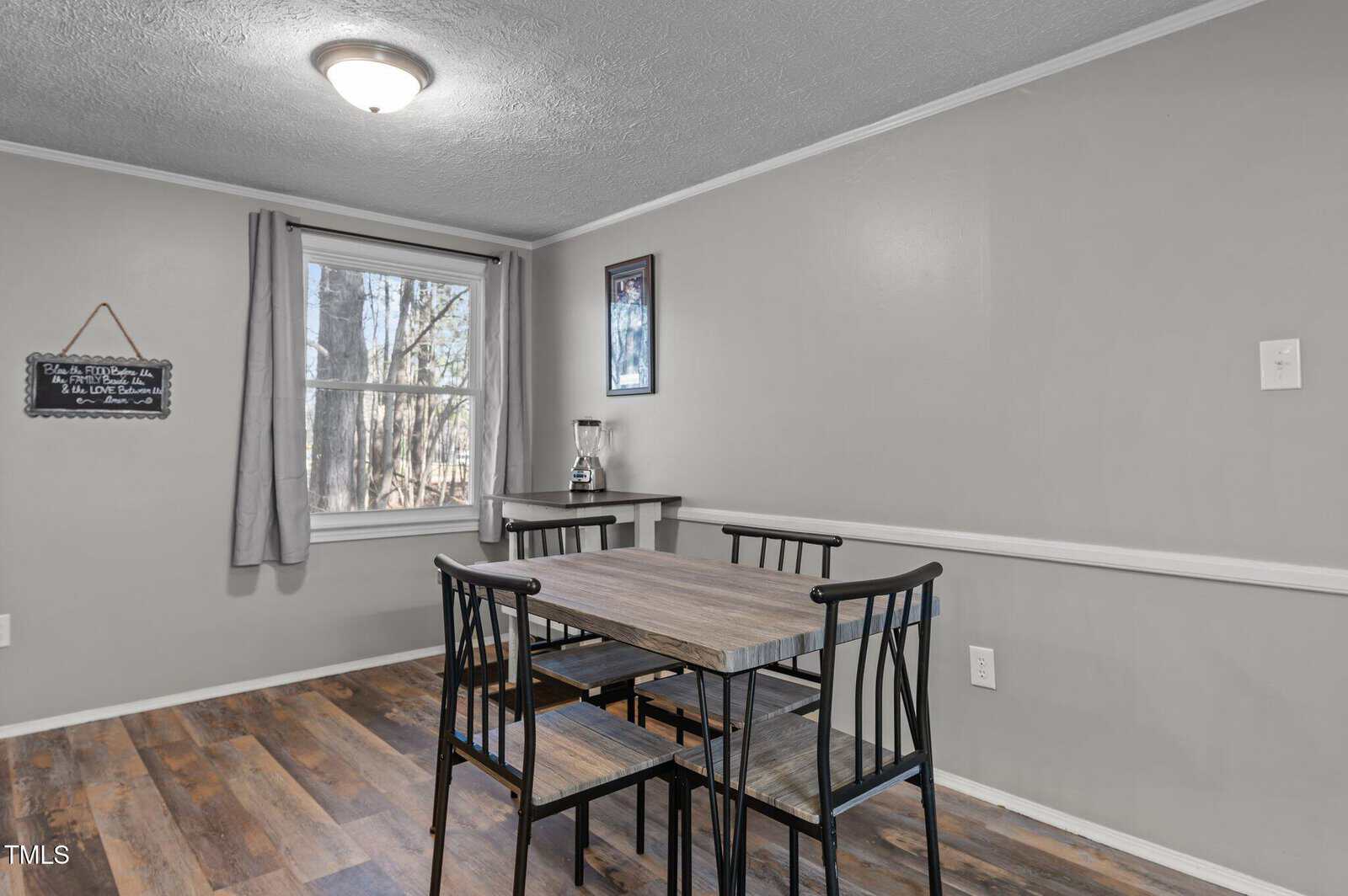 A dining area with a rectangular wooden table and four black chairs on a wood-look floor. A window with gray curtains is on the left, and a chalkboard hangs on the wall. A blender sits on a small table under a framed picture.