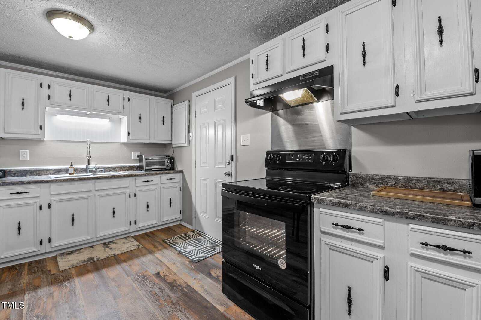 A kitchen with white cabinets and a black stove. The countertops are dark gray with a speckled pattern. There is a window above the sink on the left. A small rug lies on the wooden floor near the stove and a cutting board is on the counter.