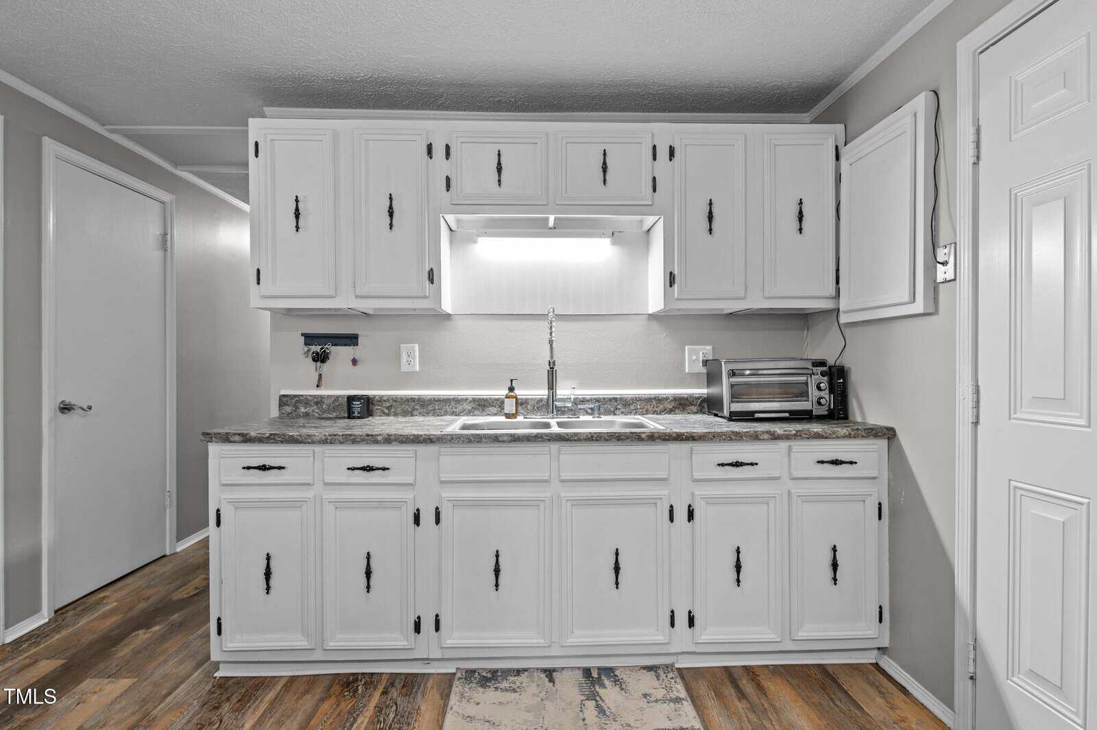A kitchen with white cabinets and a gray countertop. There is a double sink below a bright light fixture, a small toaster oven to the right, and a soap dispenser on the countertop. The floor has a wood-like pattern.