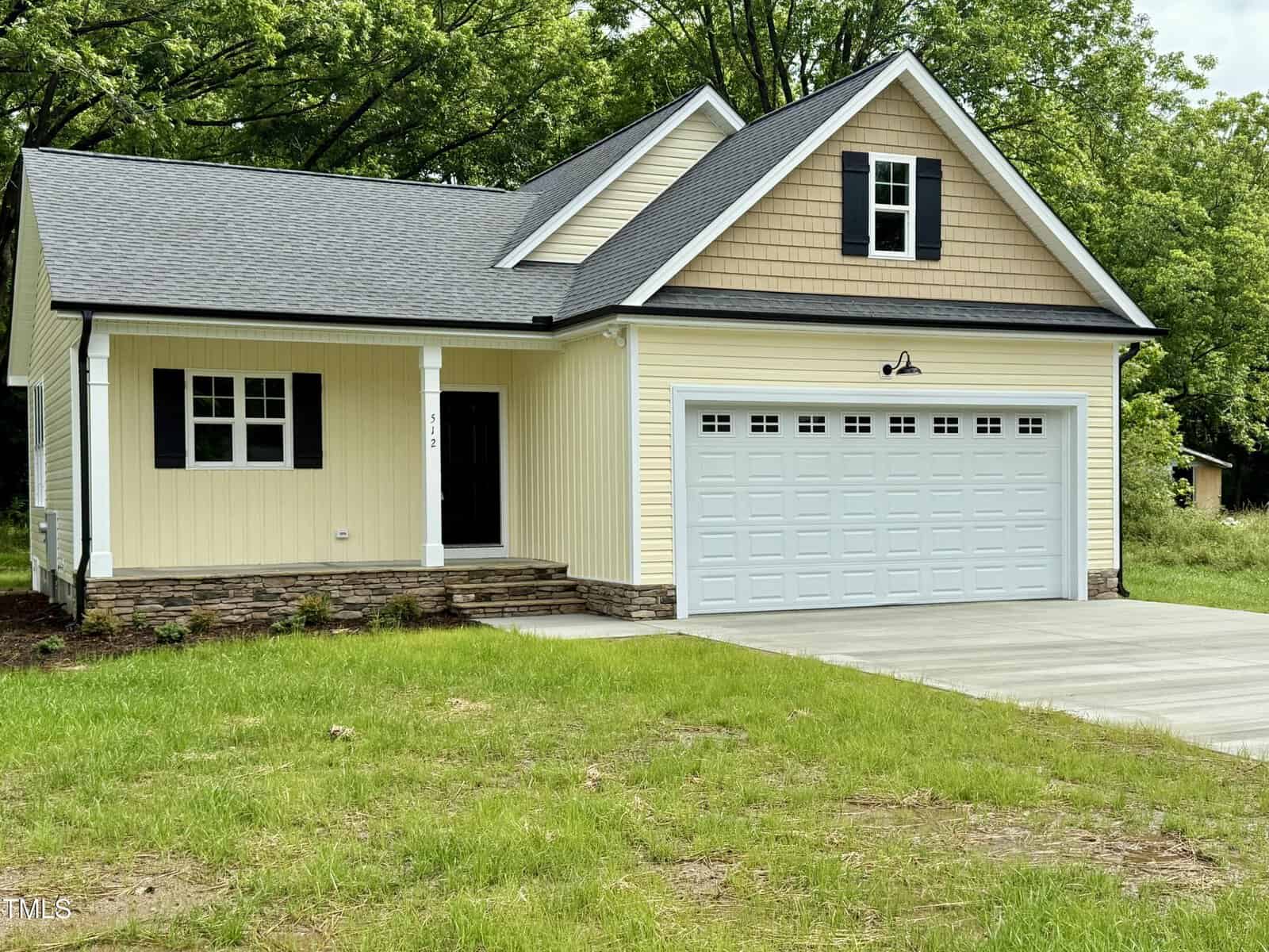 A single-story yellow house with white trim at 512 Hicksmill Road, Oxford, features a covered front porch, double garage with white doors, stone accents near the foundation, and a concrete driveway surrounded by grass and trees.