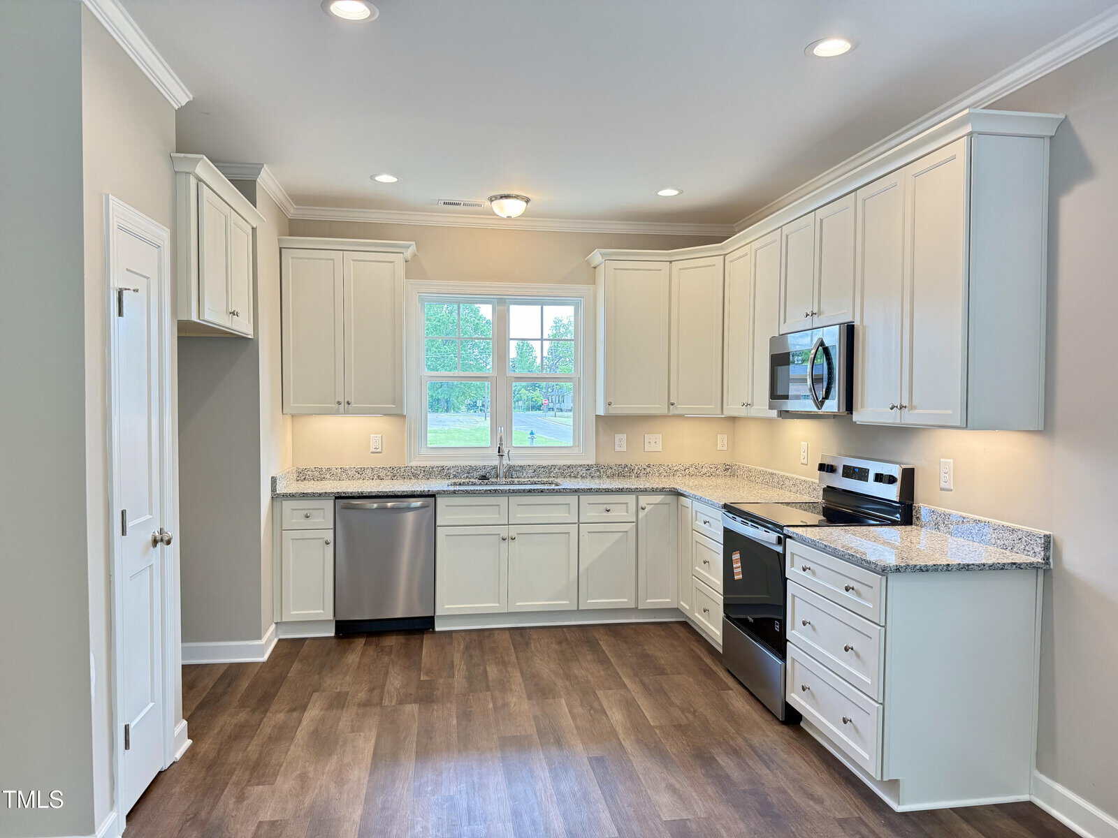 A modern kitchen with white cabinets, stainless steel appliances, granite countertops, a dishwasher, and a window above the sink. The floor is wood, and the walls are painted a light color. The room is well-lit with recessed lighting.