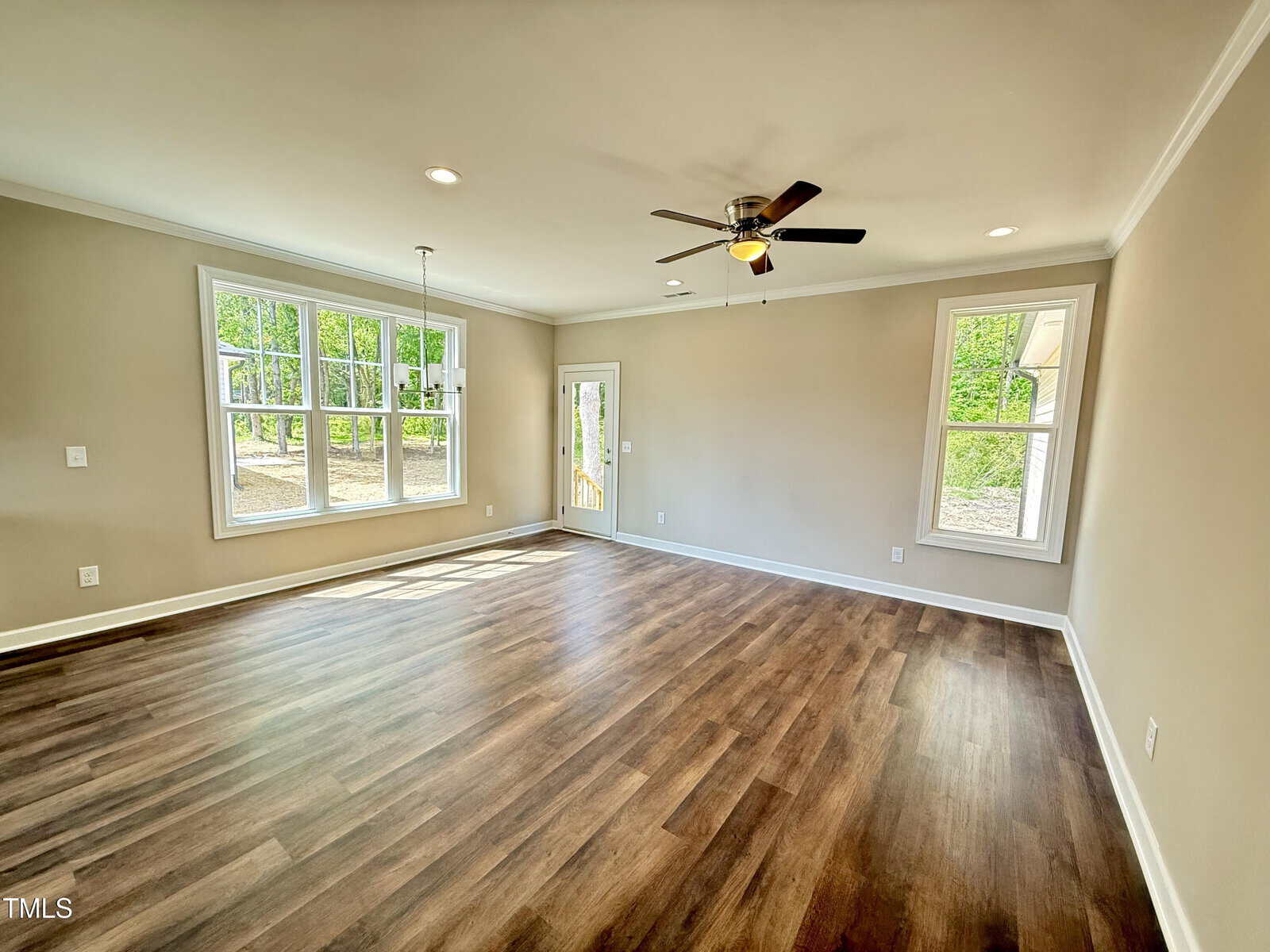 Bright, empty living room with beige walls, wood flooring, large windows, a ceiling fan with lights, and a glass door leading outside. Natural light fills the space and trees are visible through the windows.
