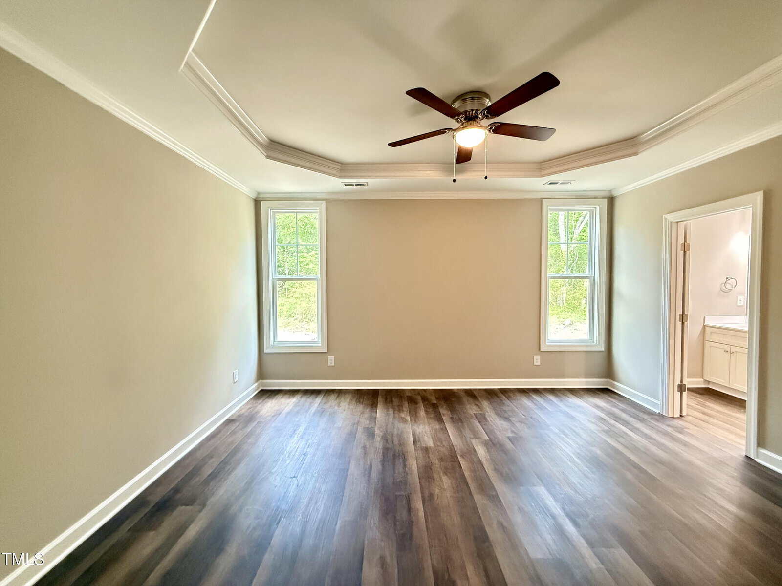 Empty room with beige walls, dark wood flooring, a ceiling fan with lights, two windows on either side of the wall, and a doorway leading to a bathroom with a visible vanity and sink.
